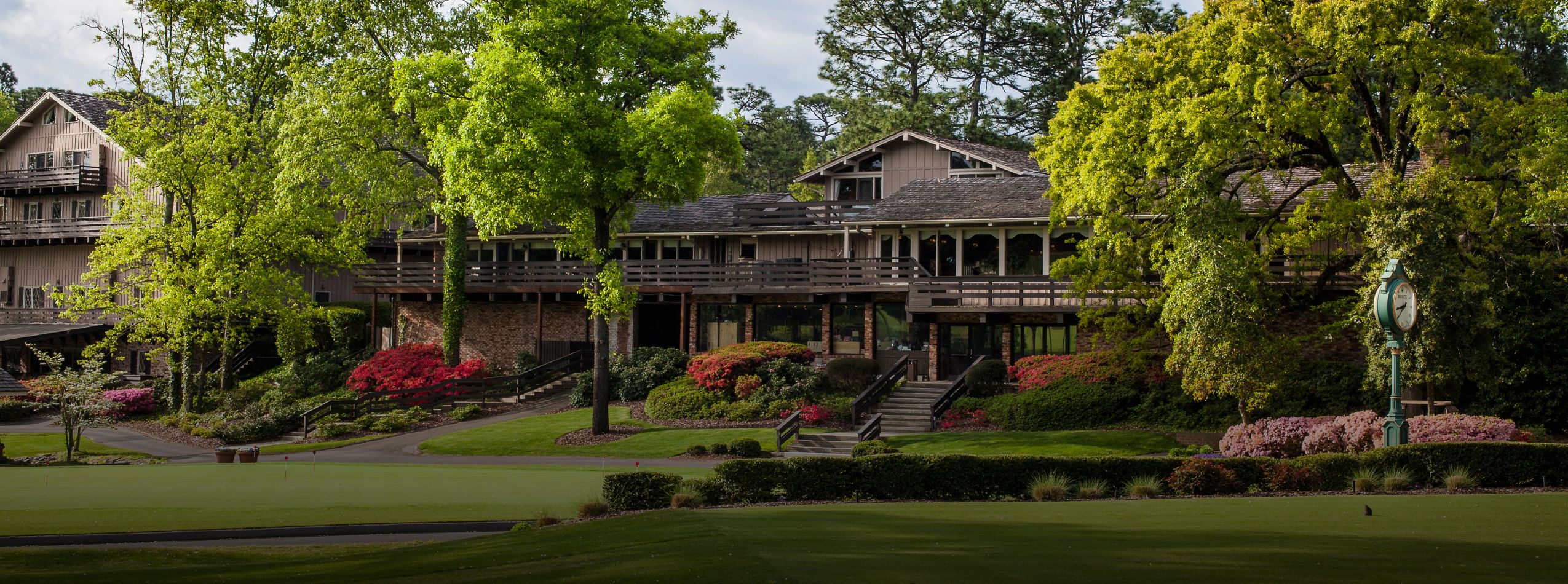 A large rustic building surrounded by lush greenery, colorful flowering plants, and trees. There are multiple levels of balconies and steps leading up to the entrance, set against a backdrop of bright blue sky and a manicured lawn in the foreground.