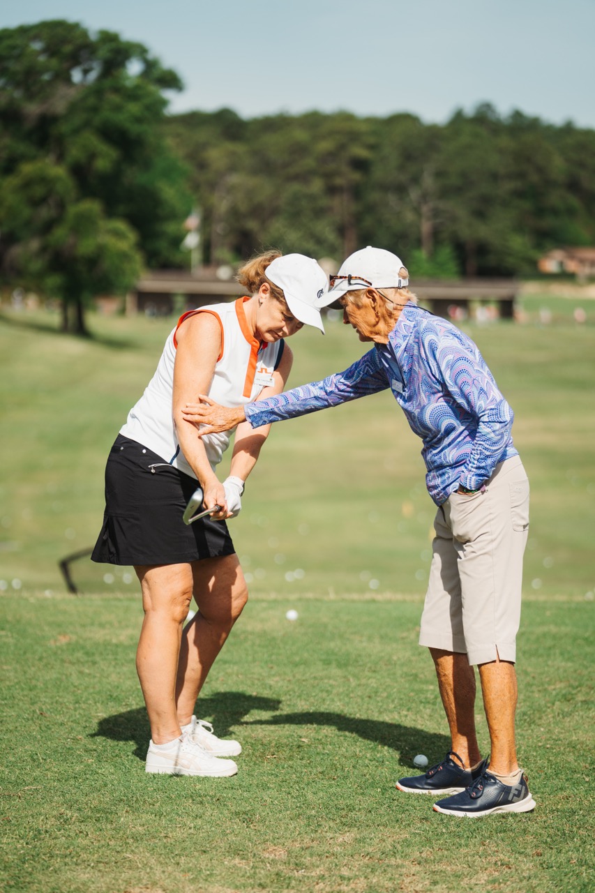 A golf instructor, wearing a blue long-sleeve shirt and light trousers, is demonstrating a technique to a student in a white sleeveless shirt and black skirt. They are on a green golf course with trees in the background.