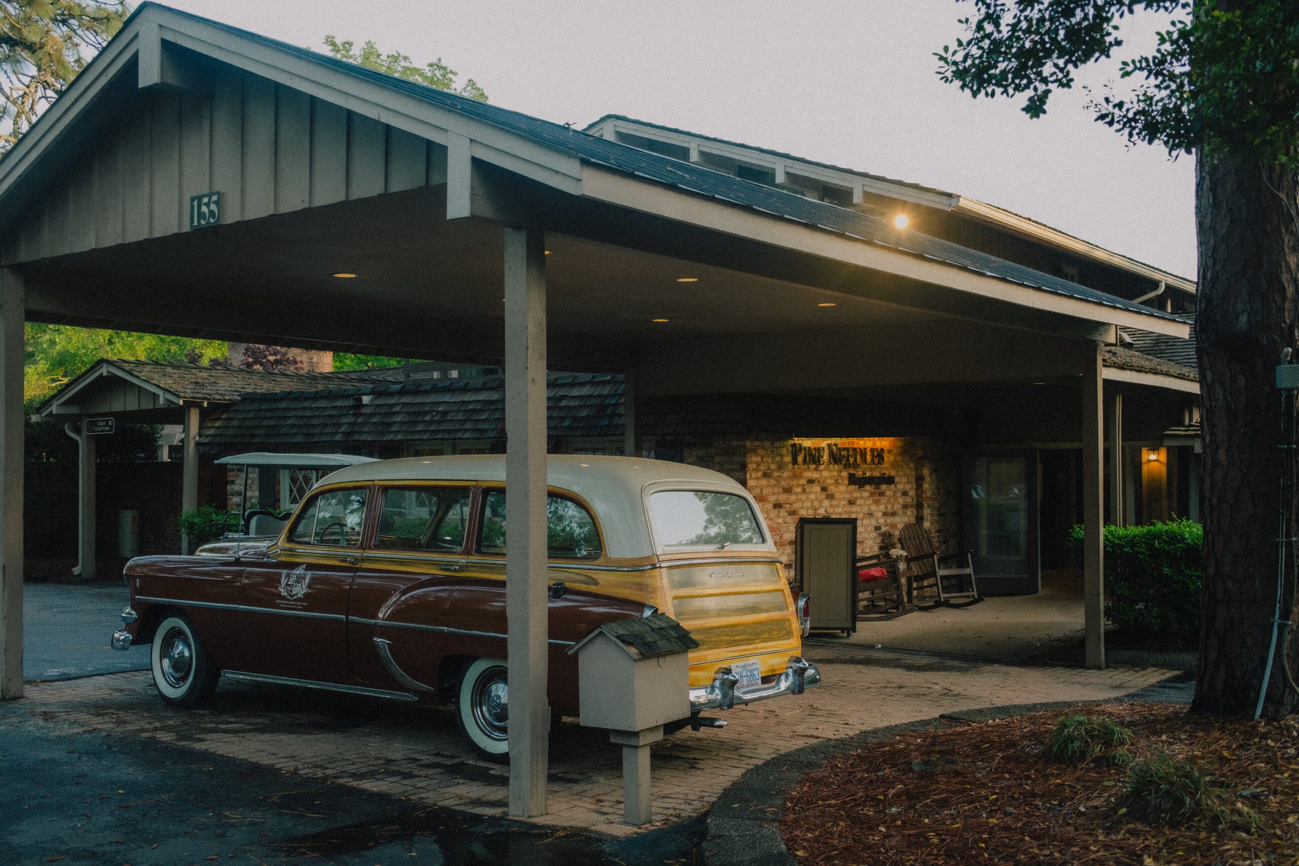 A vintage car parked in front of a building with a wooden overhang. The car is primarily light brown with yellow accents. The building's stone facade is visible, along with a sign reading "The Inn." Trees and seating areas are in the surroundings.