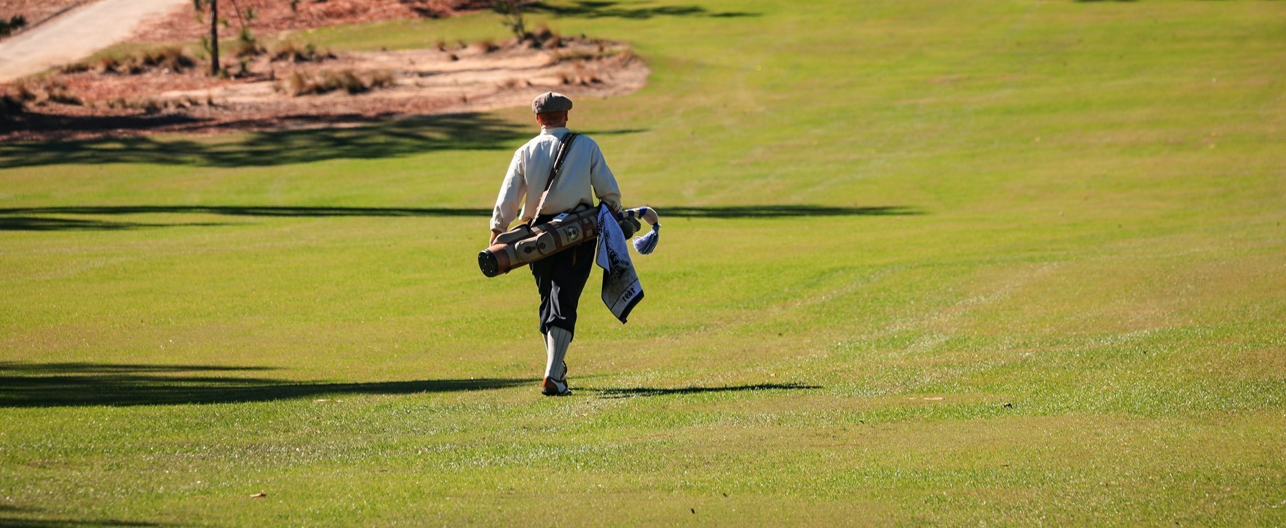 A golfer walking away from the camera on a well-maintained green golf course, carrying a golf bag over one shoulder and a blue towel in the other hand. The landscape features well-trimmed grass and a dirt path in the background.