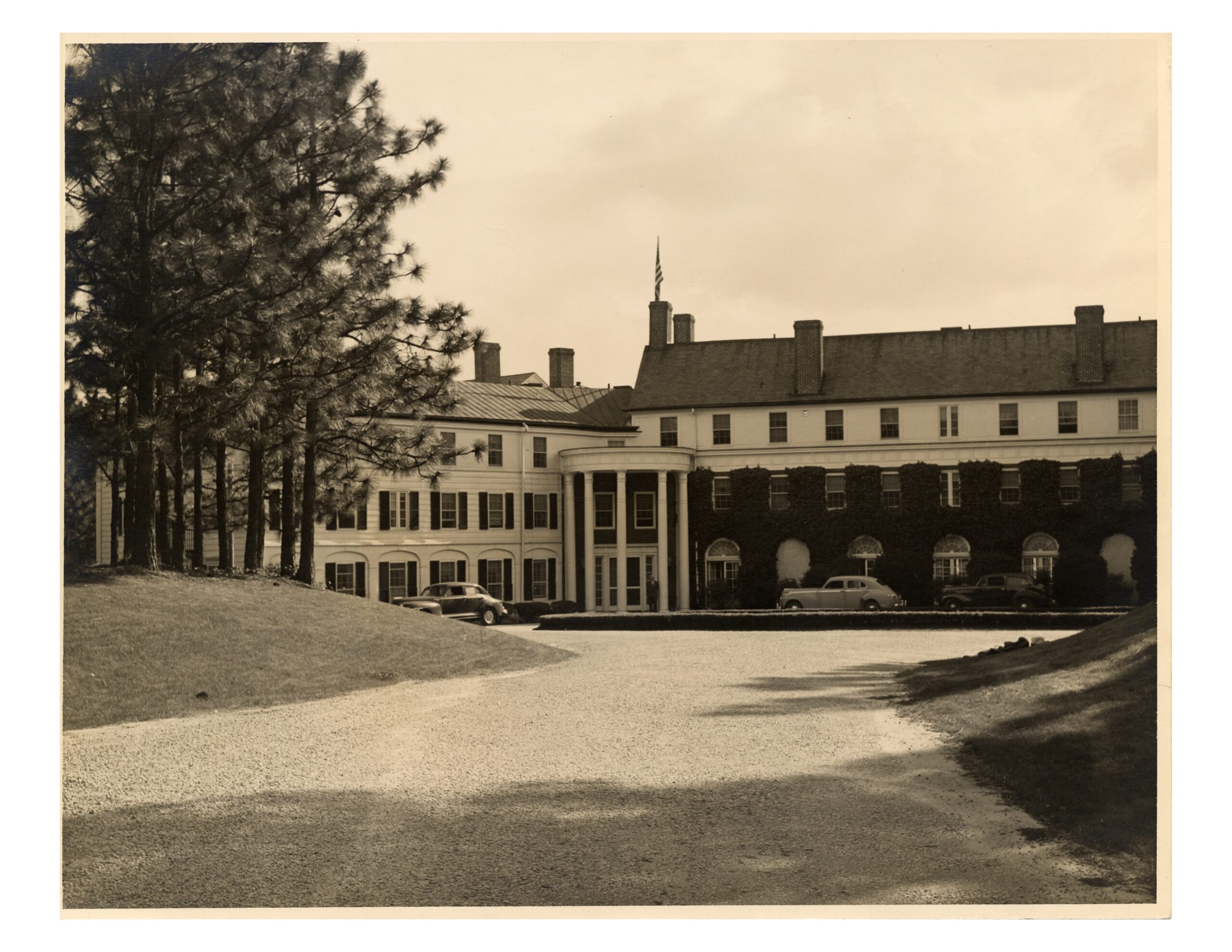A vintage black-and-white photo of a large, two-story building featuring a central entrance with columns. The front is landscaped with trees and a gravel driveway leads up to the entrance. Classic cars are parked nearby.