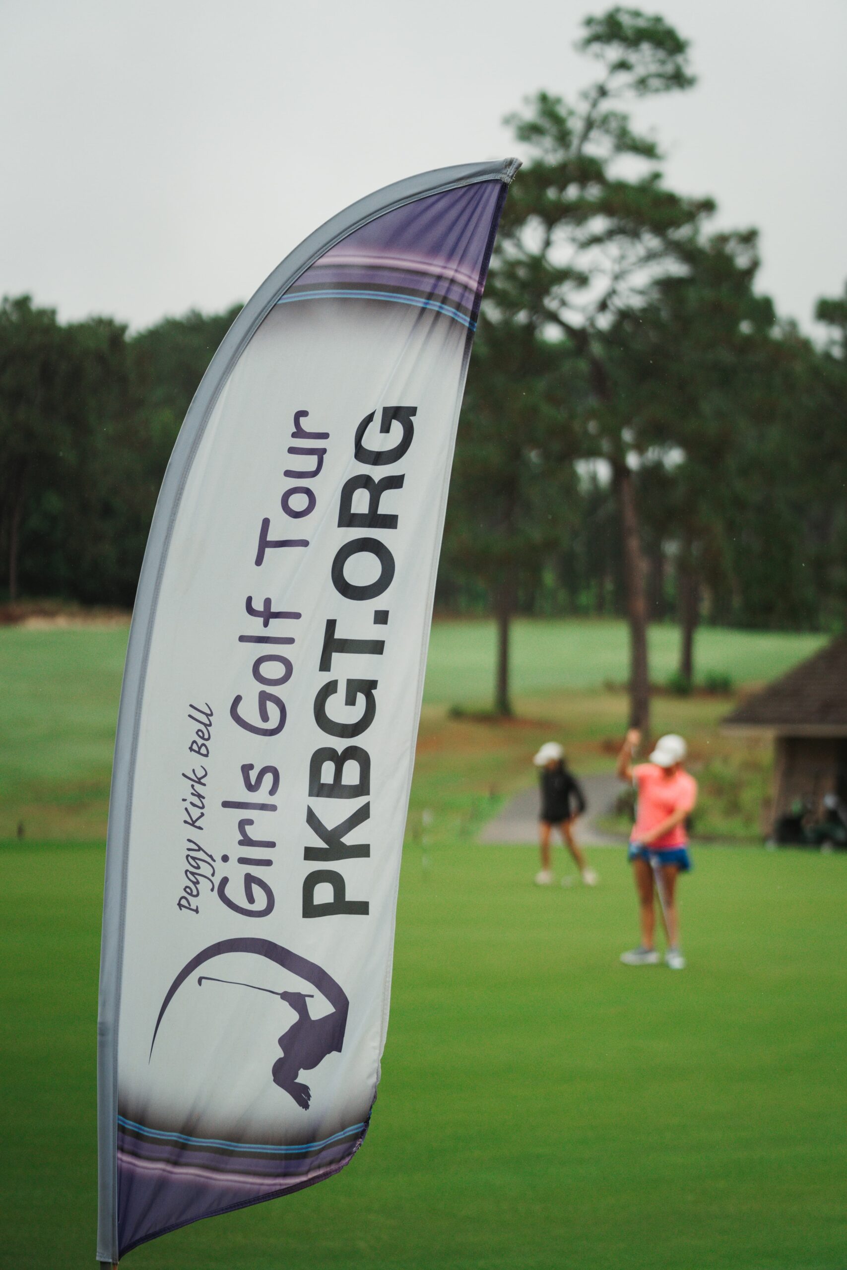 A banner for the Peggy Kirk Bell Girls Golf Tour is displayed on a golf course. The banner features the text 'Girls Golf Tour PKBGT.ORG' with a graphic of a golfer. In the background, two golfers are seen preparing to play.