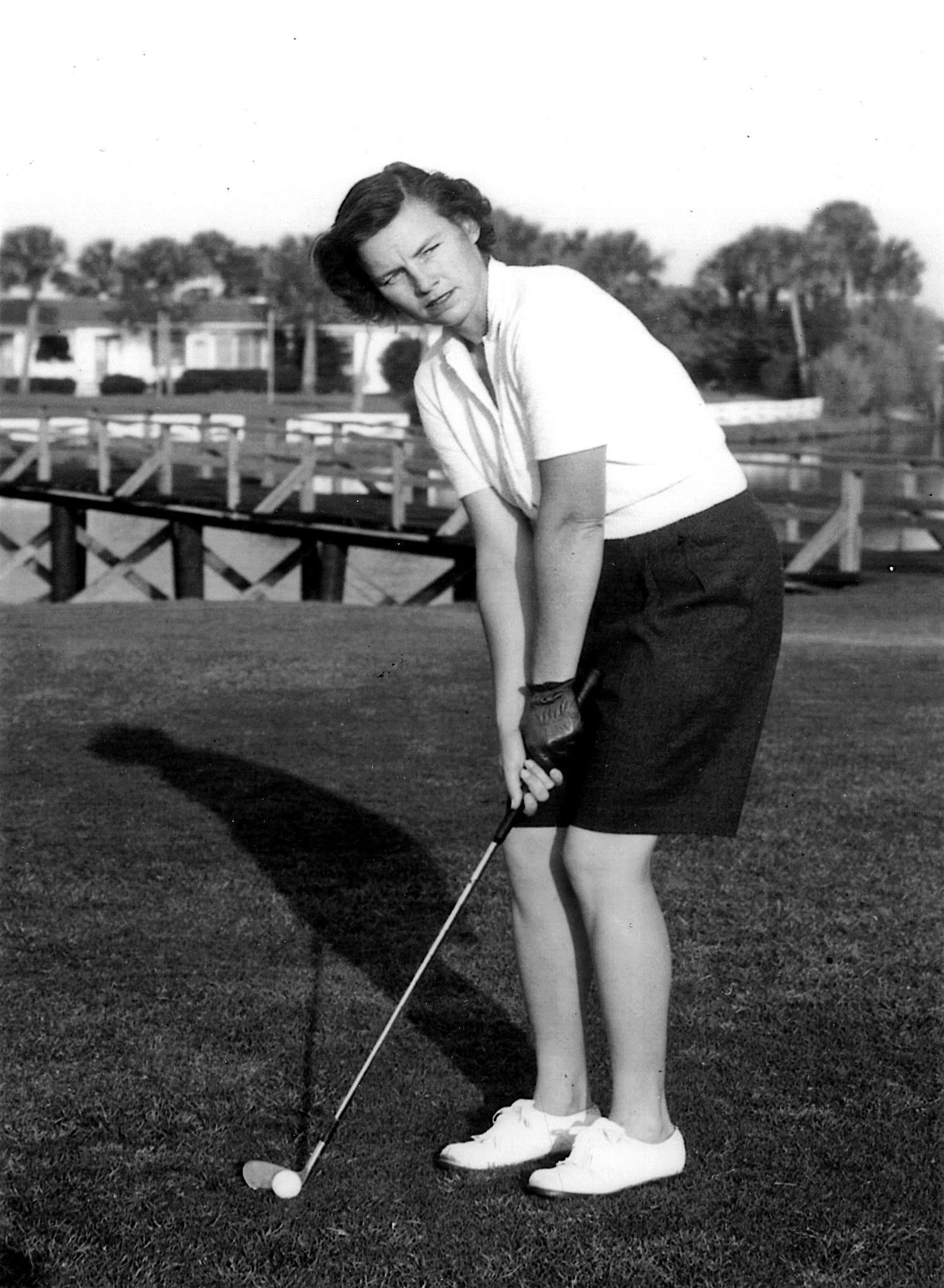 A young woman wearing a white polo shirt and dark shorts is preparing to putt on a golf course. She stands with her knees slightly bent, holding a golf club, with a focus on her stance. The background shows a wooden structure near the course.