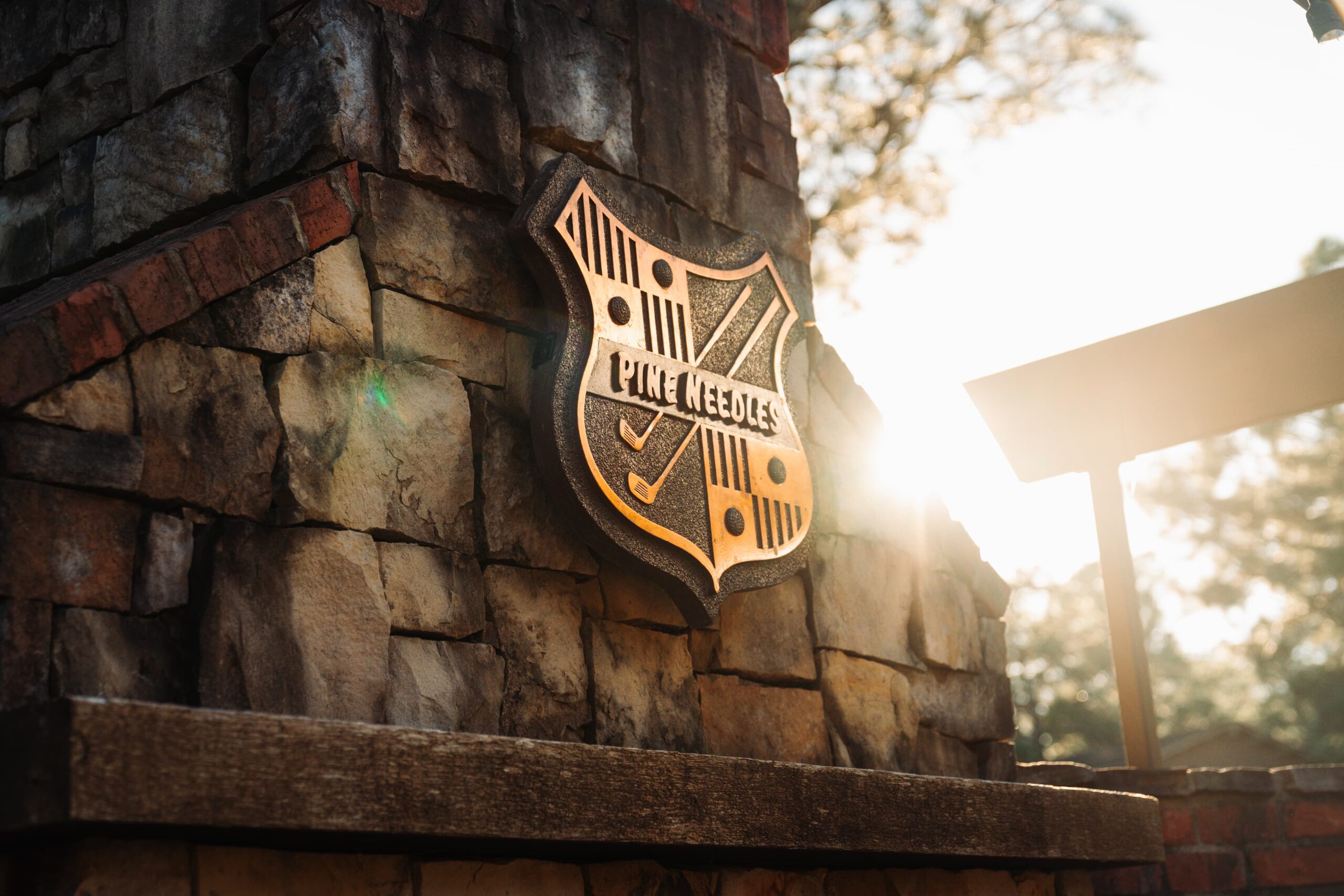 A close-up of a stone monument featuring a shield-shaped logo with the text "Pine Needles" and a golf club graphic. The sun is shining softly in the background, illuminating the stone surface.