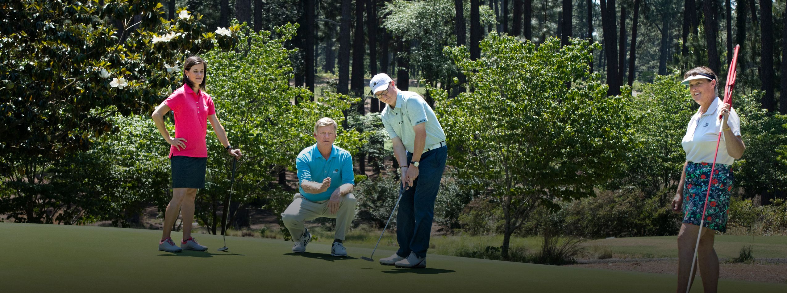 Four individuals are on a golf course, focused on a putting green surrounded by trees. Two men, one in a blue shirt and one in white, are crouched over the ball. Two women stand nearby, one in a pink shirt and the other in a patterned skirt holding a flag.