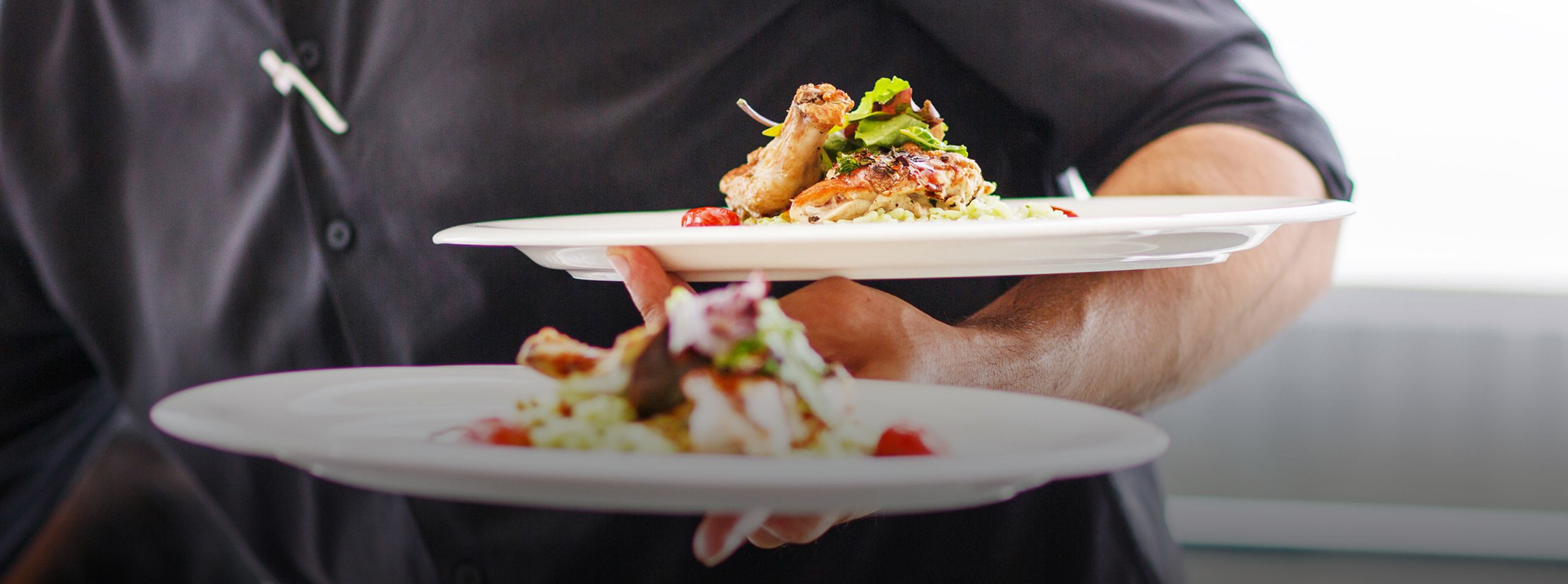 A person in a black shirt holding two white plates with food. The upper plate features a piece of grilled chicken garnished with greens, while the lower plate contains a salad with mixed ingredients and small red accents.
