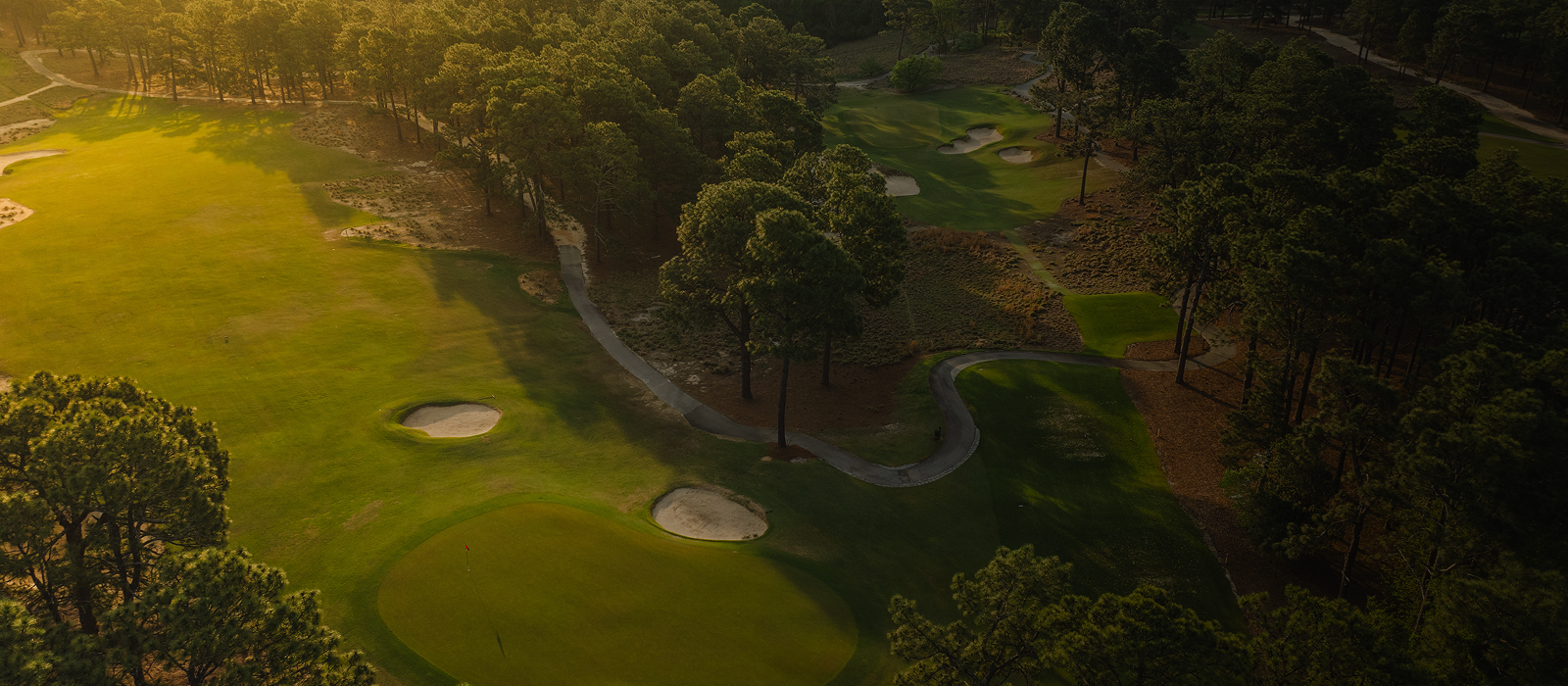 Aerial view of a golf course featuring manicured fairways, sand traps, and green areas surrounded by trees. The scene is illuminated by soft golden sunlight, highlighting the contours of the landscape.