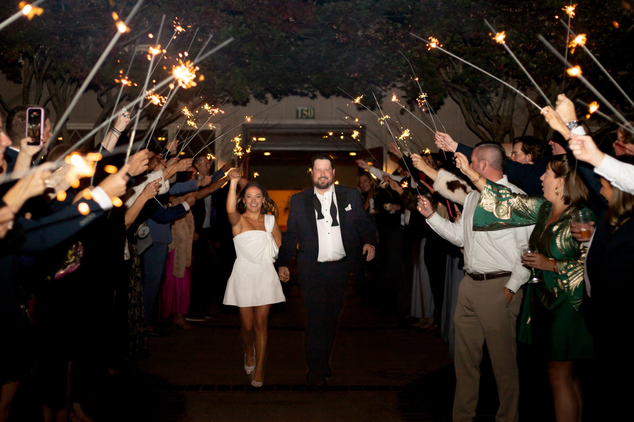 A bride in a white dress and a groom in a black tuxedo walk hand in hand under a canopy of sparklers held by guests at an outdoor celebration. The scene is illuminated by the sparkler light, creating a festive atmosphere.