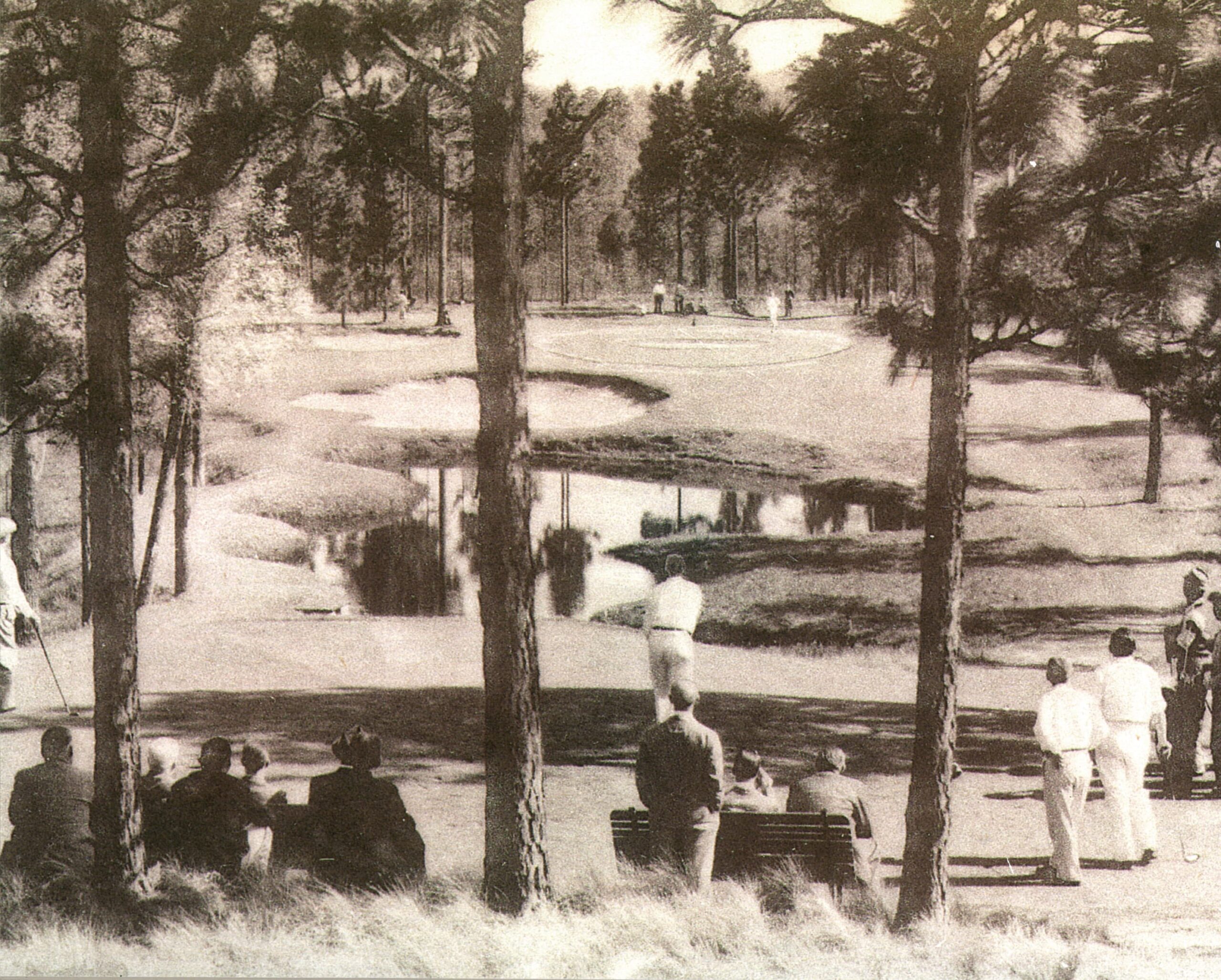 A black and white historical photo of a golf course, showcasing a player preparing to putt. Spectators sit on benches in the foreground, while trees frame the scene. The course features a winding water hazard and neatly maintained grass.