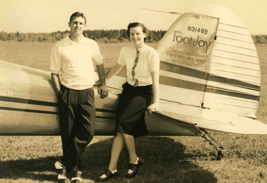 A man and a woman pose next to a small plane on a grassy field. The man wears a light shirt and dark trousers, while the woman is dressed in a blouse and skirt, smiling. The plane features 'FootJoy' on its side and is parked against a tree-lined background.
