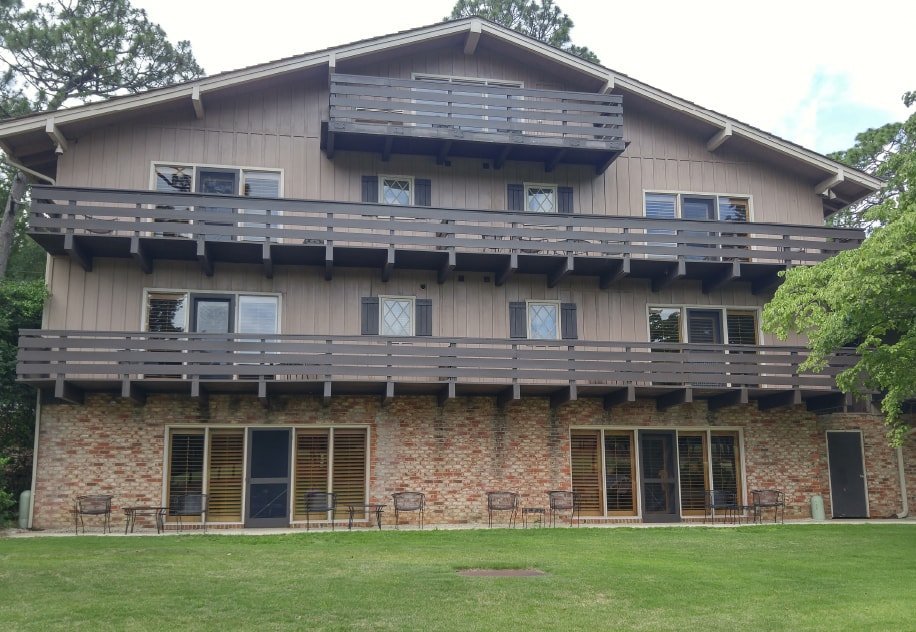 A multi-story building with a brown wooden exterior and brick lower section, featuring multiple balconies on the upper floors. The ground floor has large glass doors, leading to a grassy area with patio chairs in front.