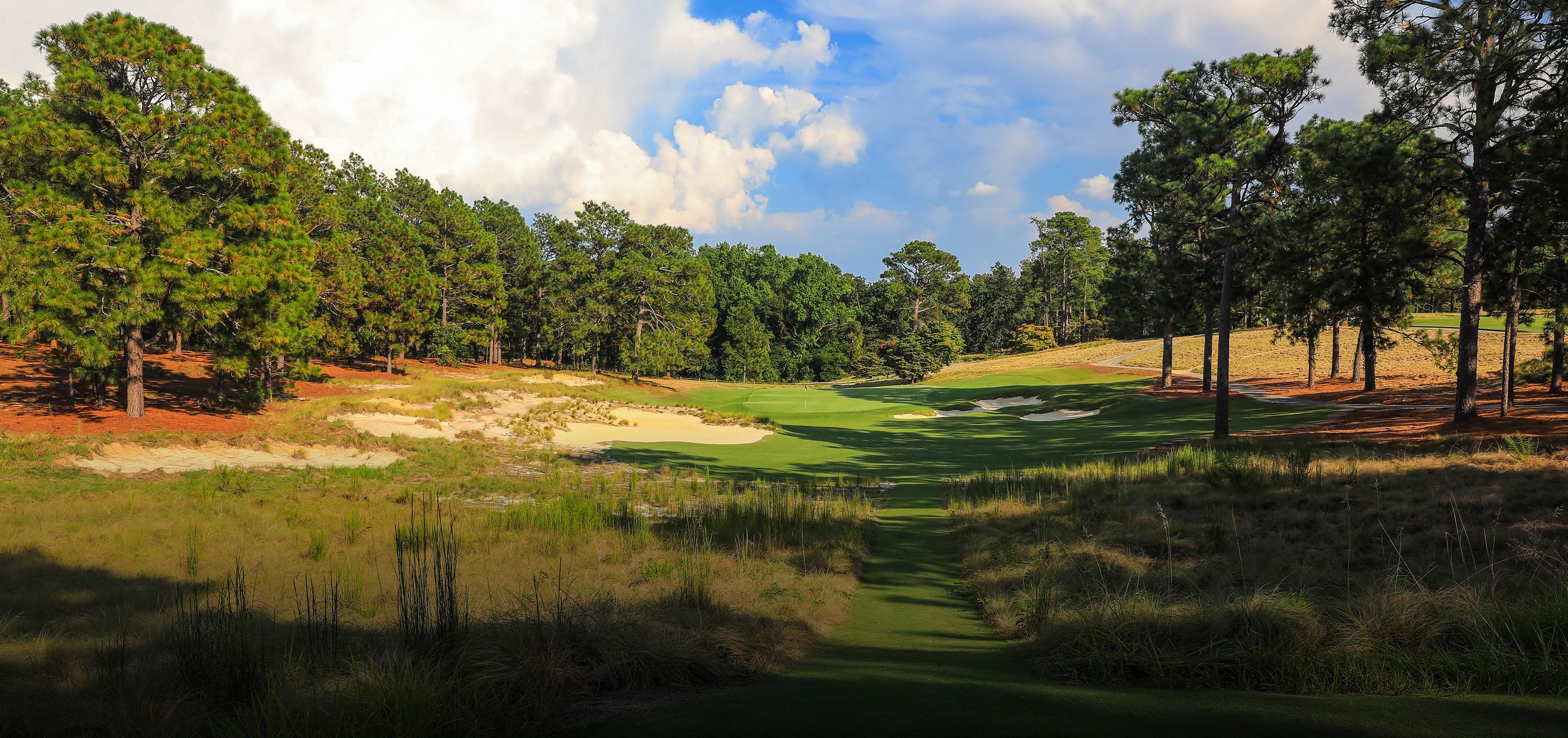 A scenic view of a golf course fairway lined with green grass and clusters of trees on either side. The sky is partly cloudy, and the landscape features sandy patches in the background, creating a blend of natural elements in a serene environment.