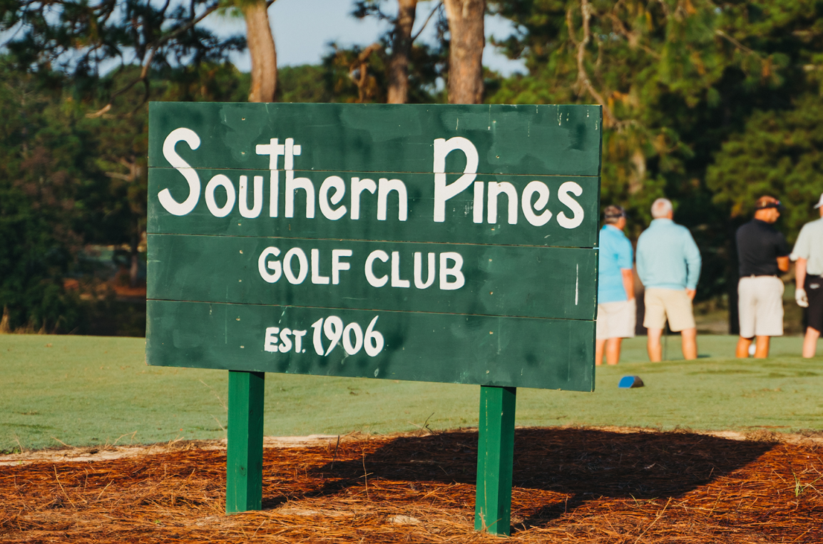 A wooden green sign reading "Southern Pines GOLF CLUB EST. 1906" stands in front of a golf course, with several people in the background engaged in a golf activity. The setting features trees and grass typical of a golf course.