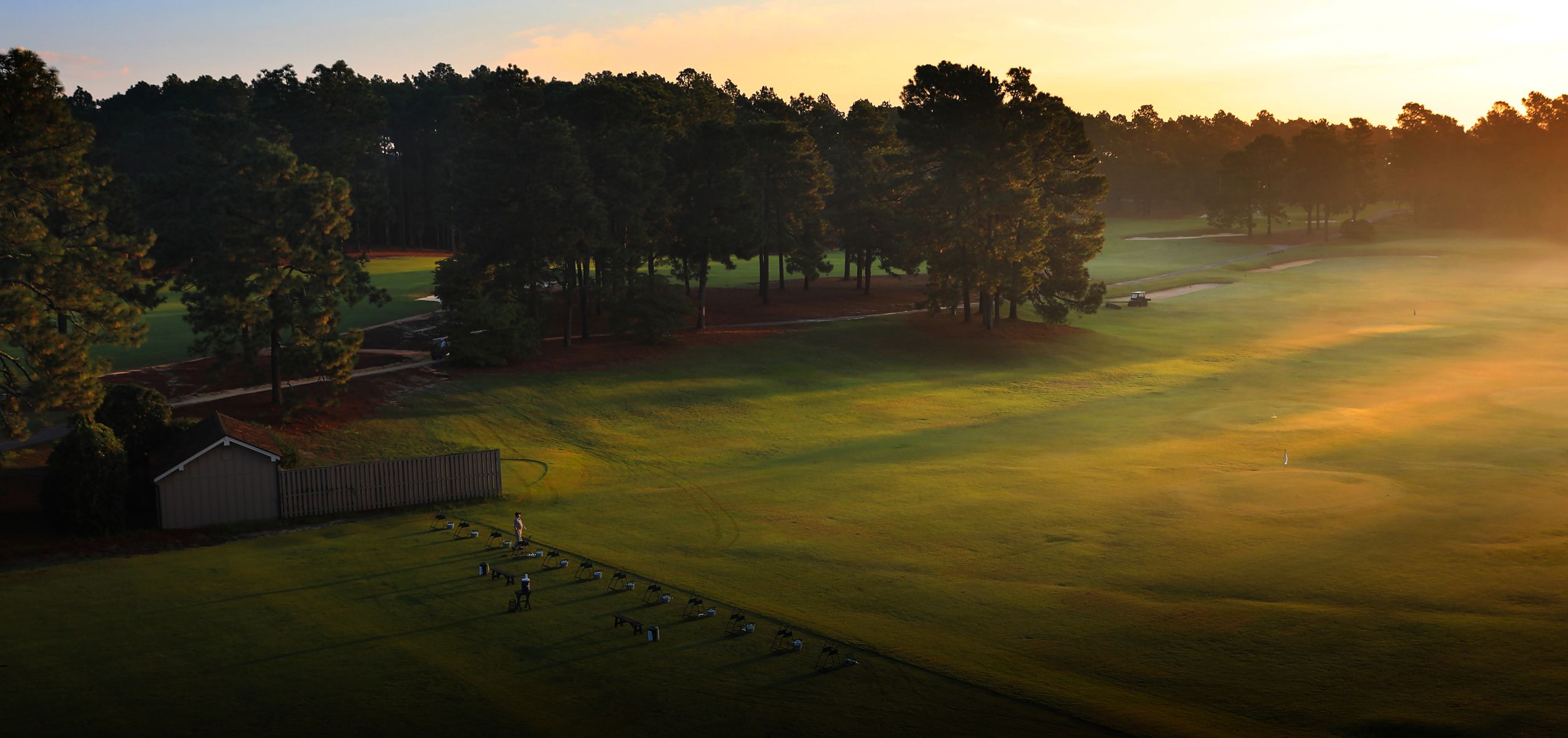 A scenic view of a golf course at sunrise, featuring green grass, trees lining the fairways, and a misty atmosphere. The area also includes a small building and numerous golf tees positioned for practice.