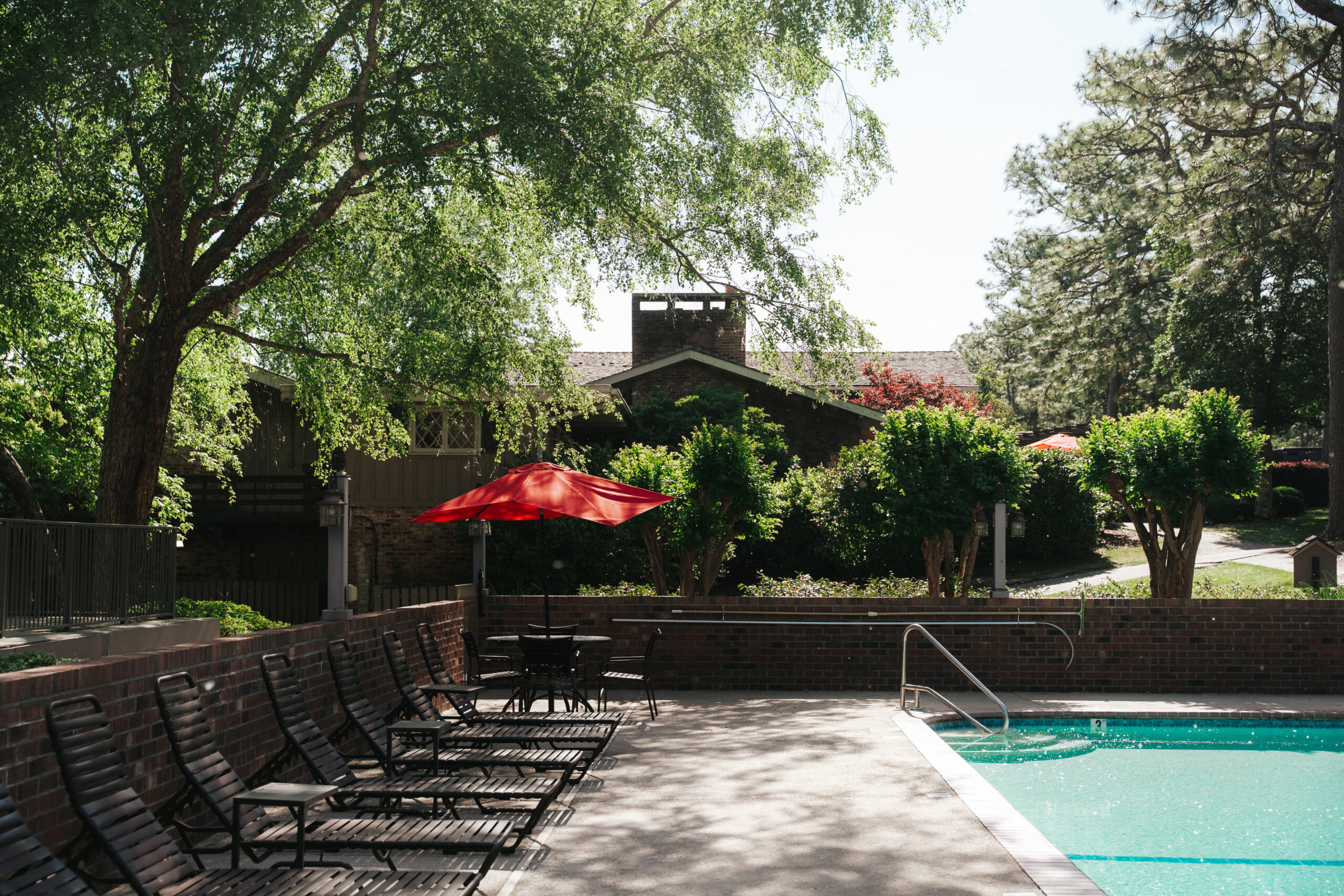 A swimming pool area surrounded by lounge chairs under the shade of trees. An umbrella in bright red color is placed over a small table. In the background, there are buildings partially obscured by greenery, indicating a residential setting.