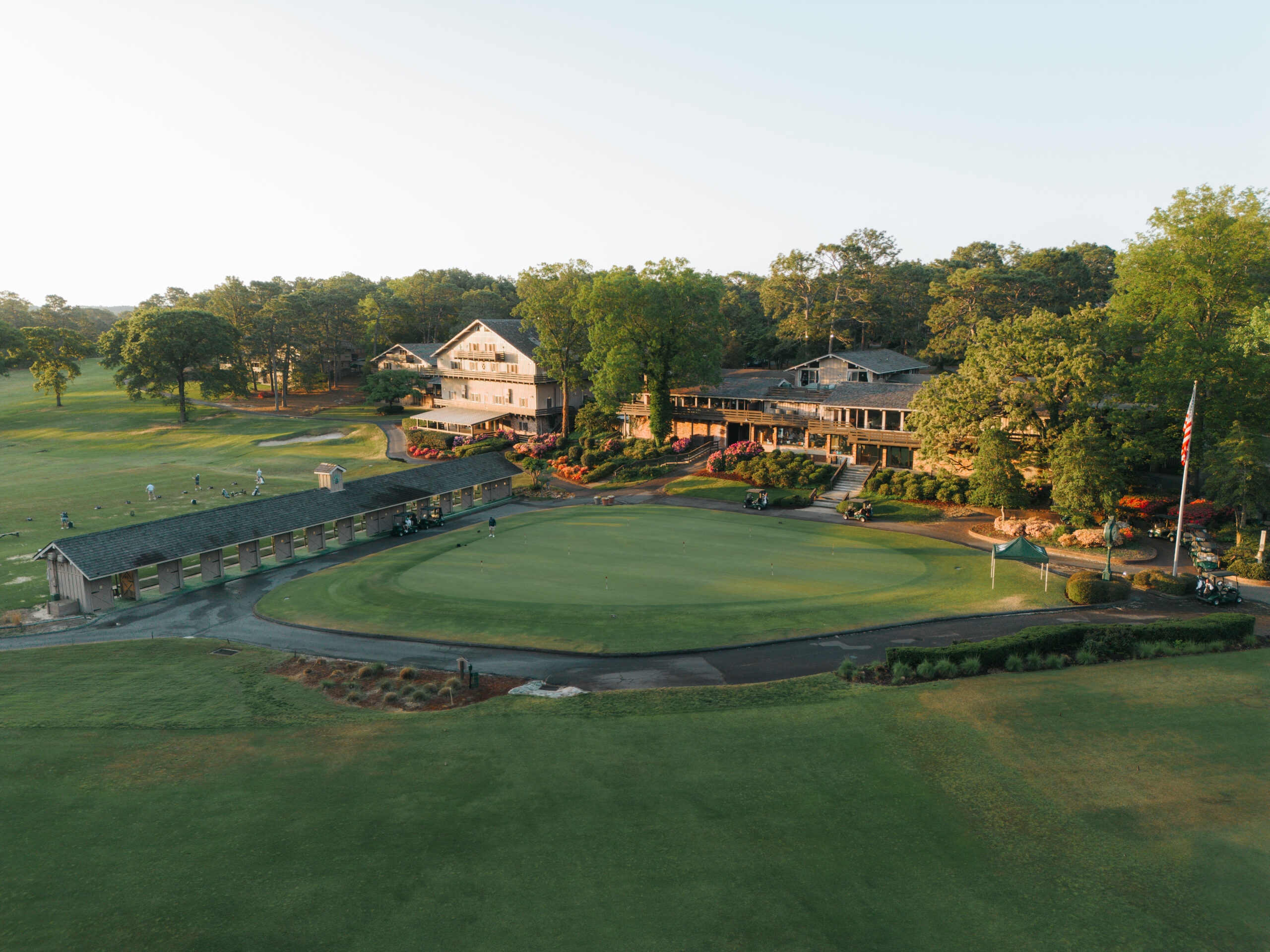 Aerial view of a golf course clubhouse surrounded by lush greenery. The clubhouse features a large porch and several windows. Nearby is a practice area with golf clubs and a putting green, with trees and fairways extending in the background.
