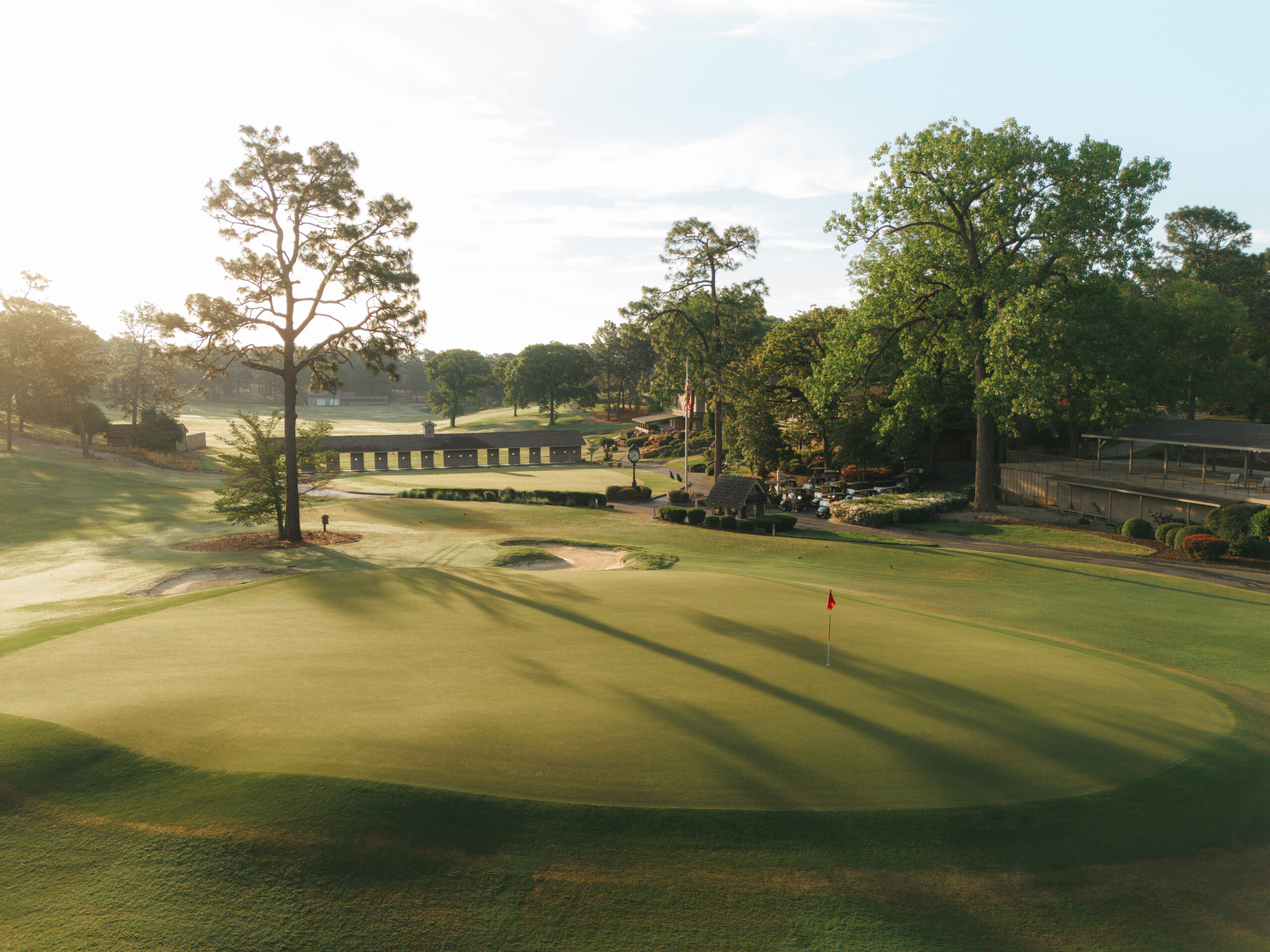 A scenic view of a golf course in the early morning, featuring a well-maintained green with a flag. Surrounding trees and a bridge are visible in the background, along with a clubhouse and parked cars.