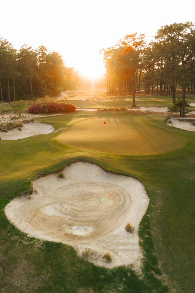 A golf course with several sand bunkers and a well-maintained green. The setting sun creates a warm glow over the landscape, highlighting trees in the background and colorful shrubs near the putting area.