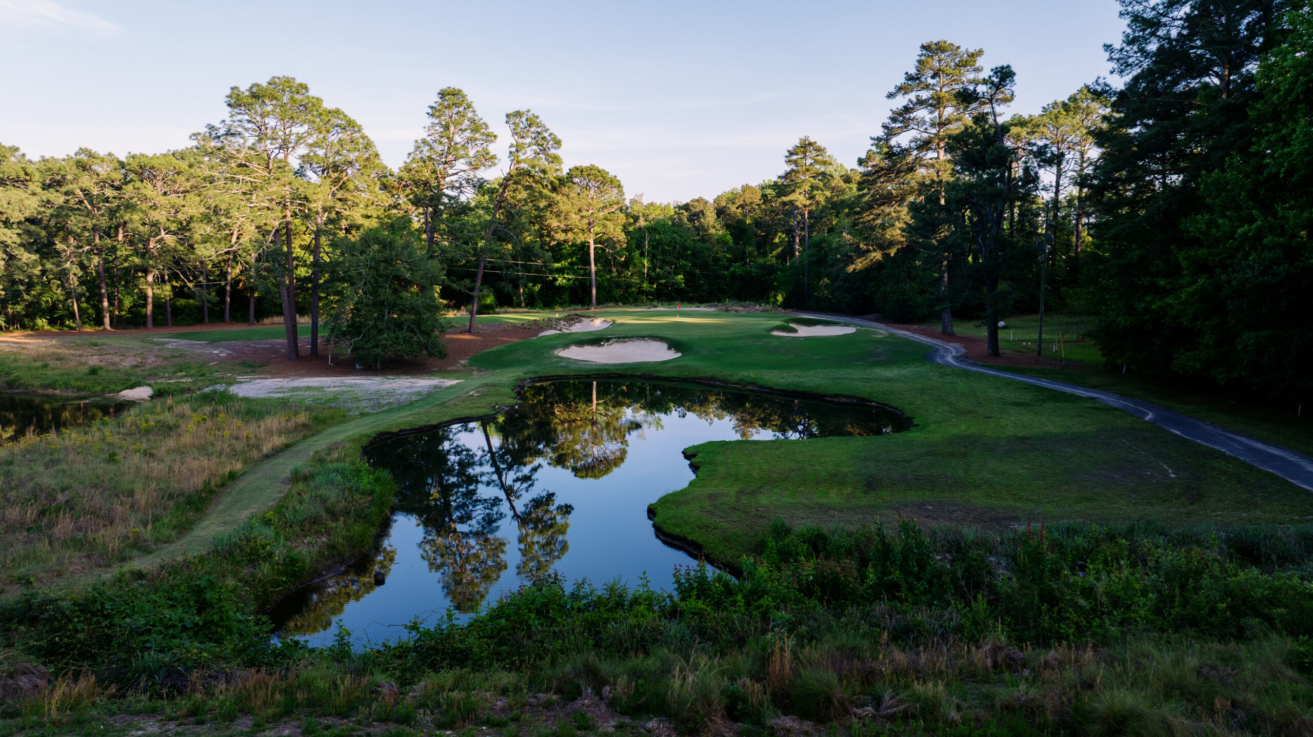 A scenic view of a golf course with a water hazard, surrounded by lush trees. The green grass features a sand bunker to the left, and a calm pond reflects the blue sky and nearby foliage. A pathway runs along the edge of the course.