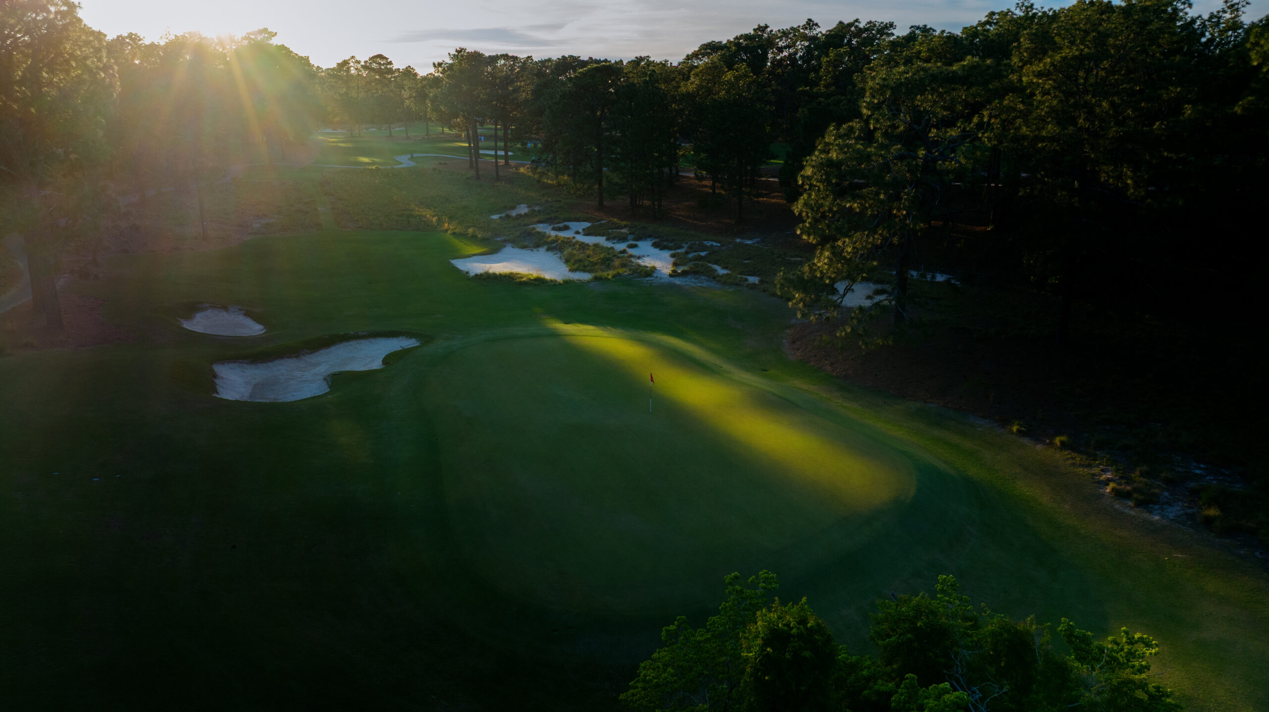 An aerial view of a golf course during sunrise or sunset, showcasing lush green fairways, sand bunkers, and scattered trees. The sun's rays create a warm glow over the landscape, illuminating the colors of the course.