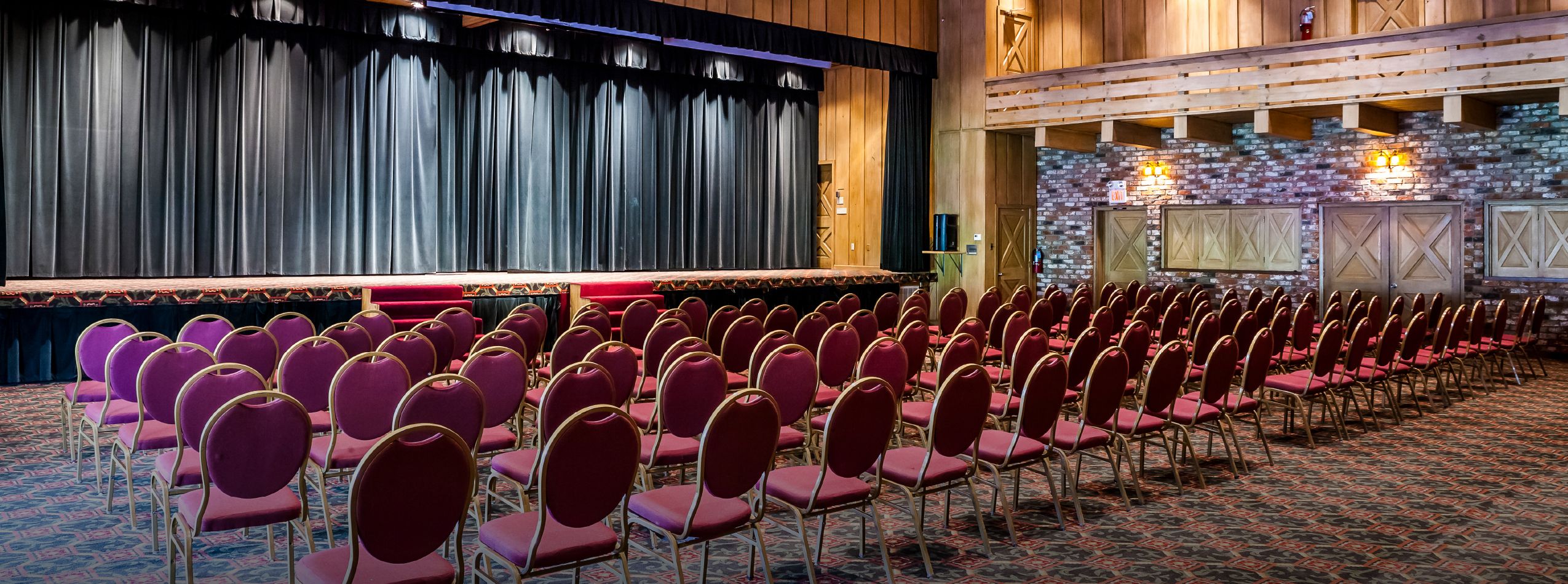 An indoor auditorium featuring rows of red upholstered chairs arranged in front of a raised stage with a black curtain. The wooden walls and decor contribute to a formal atmosphere, with lighting positioned above the stage.