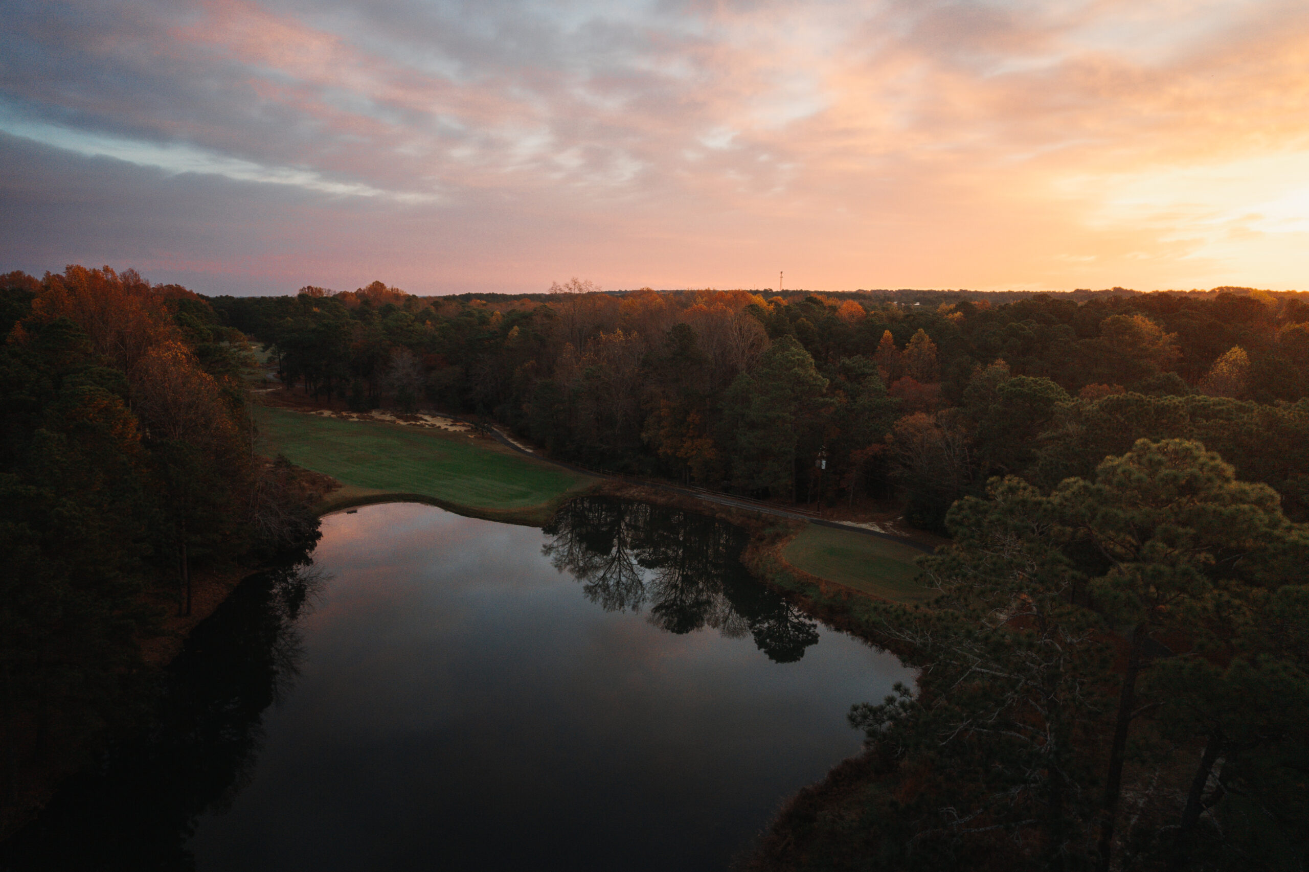 Aerial view of a serene landscape at sunset, featuring a still lake reflecting the colorful sky. Surrounding the lake are trees with autumn foliage. The scene captures a blend of greens and warm hues, creating a peaceful natural setting.