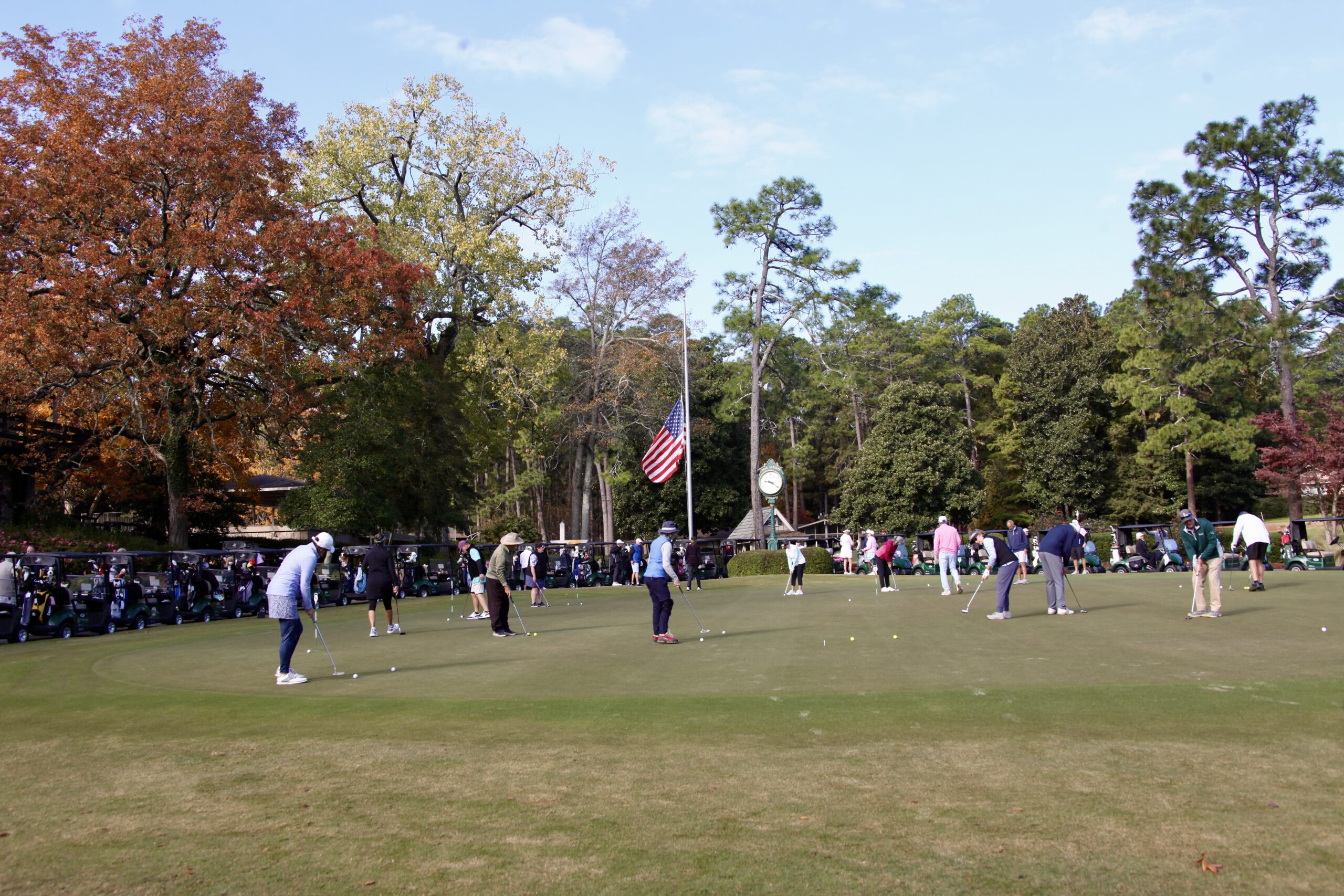 Participants practicing putting on a green with audience members seated in the background. A large American flag is displayed, and trees with autumn foliage surround the area, creating a colorful setting.