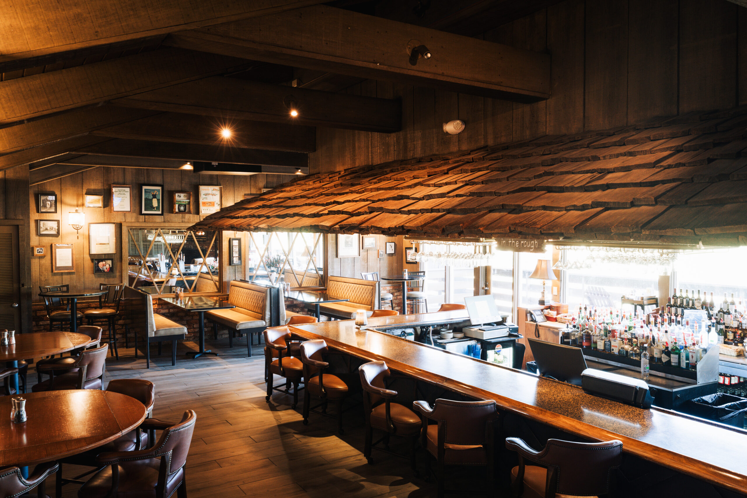 Interior of a rustic restaurant featuring wooden beams and a sloped ceiling. The bar is stocked with various bottles, and there are round tables and cushioned seating in the background, illuminated by natural light from large windows.