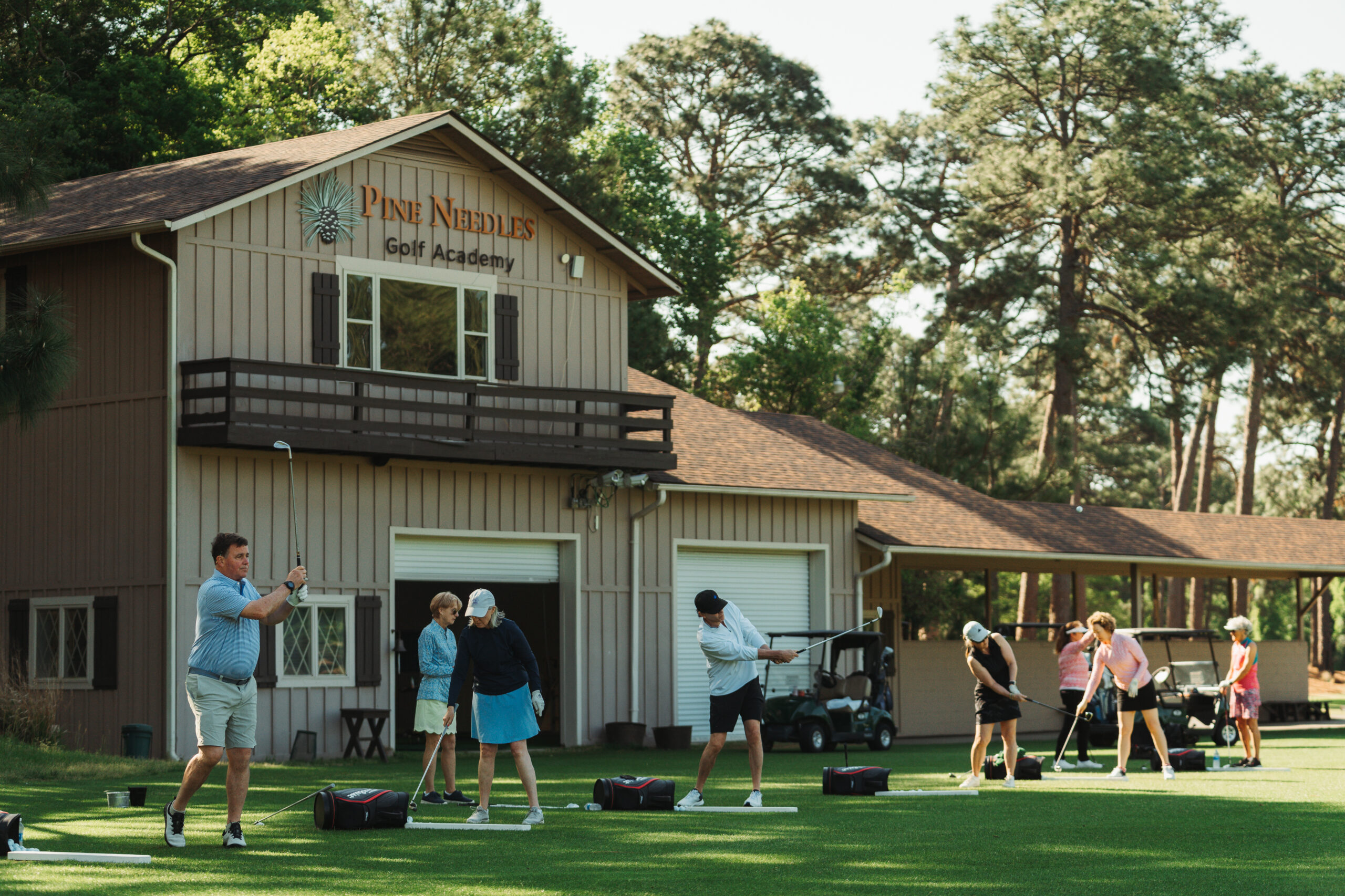 A golf academy setting with a two-story building labeled "Pine Needles Golf Academy." Multiple individuals practicing golf swings on lush green grass, while others prepare equipment nearby. Tall trees frame the background.