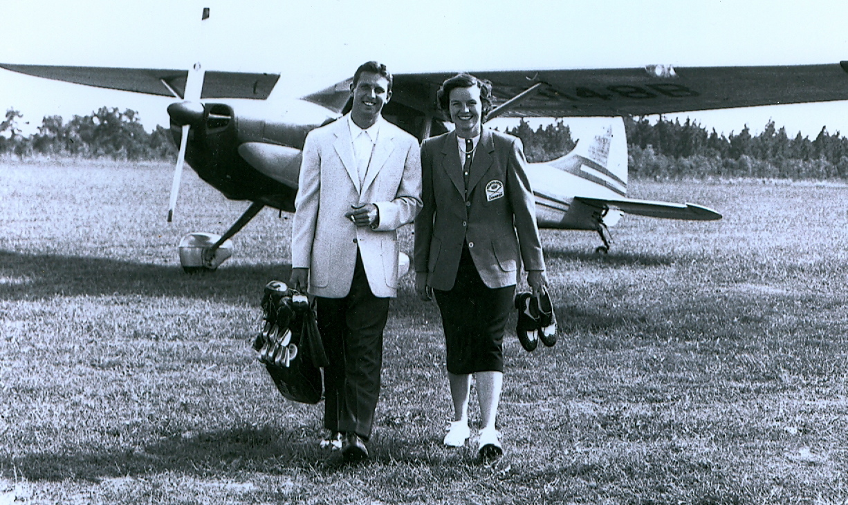 A man and a woman walking side by side on an airfield, both dressed in formal attire. The man holds a camera and the woman carries a pair of shoes. A small airplane is parked in the background, with trees visible in the distance.