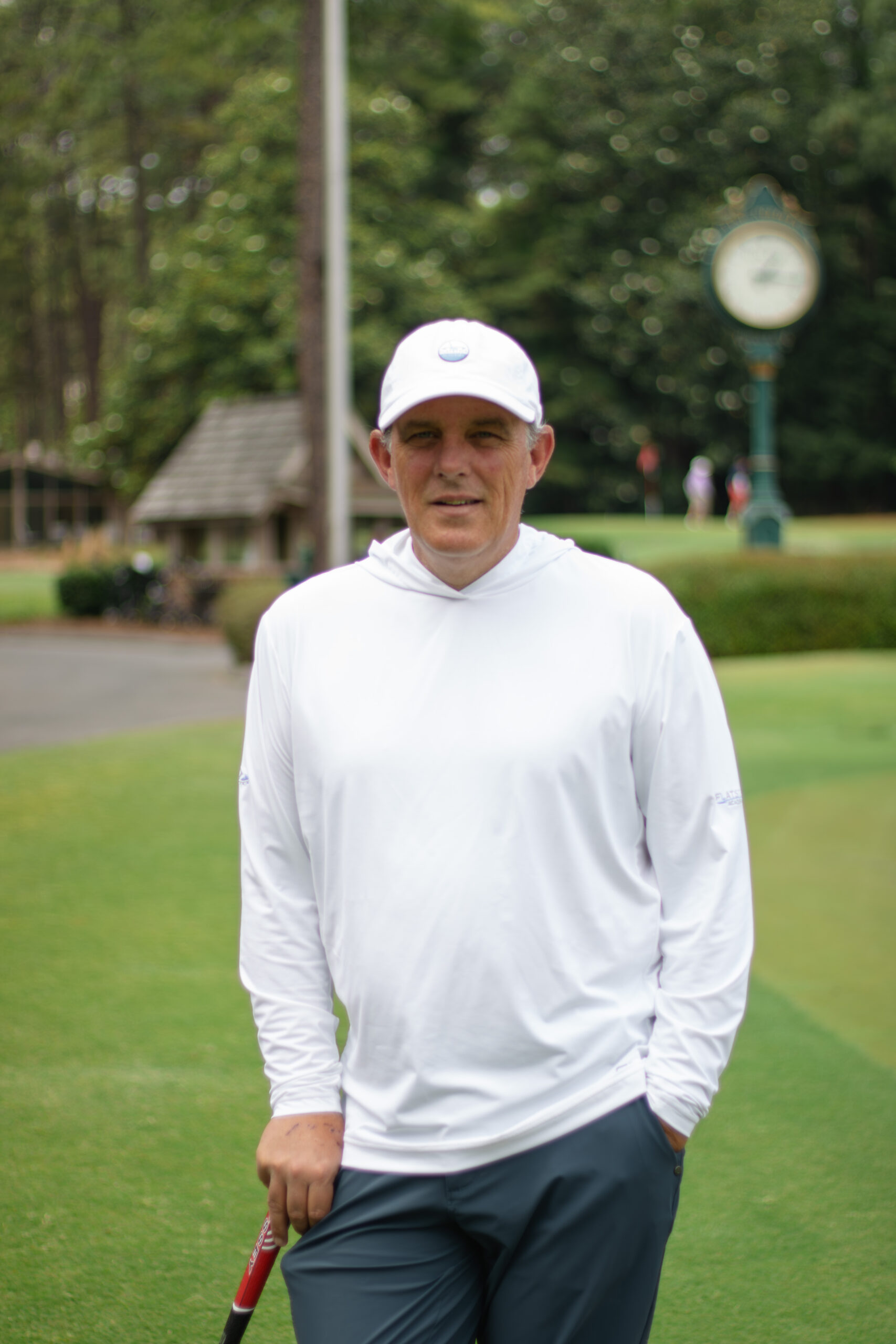 A man wearing a white hooded sweatshirt and a white cap stands on a golf course, holding a golf club. In the background, there is a clock and greenery, while other people are visible in the distance.