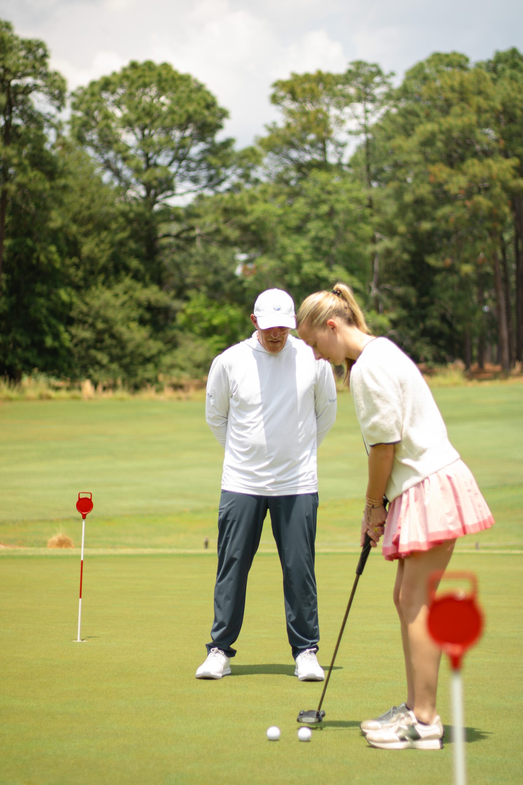 A young woman in a white sweater and pink skirt is preparing to putt on a golf green, focusing on the ball. Behind her, a man in a white cap and long-sleeved shirt watches intently. Red flags mark the putting holes in the background.