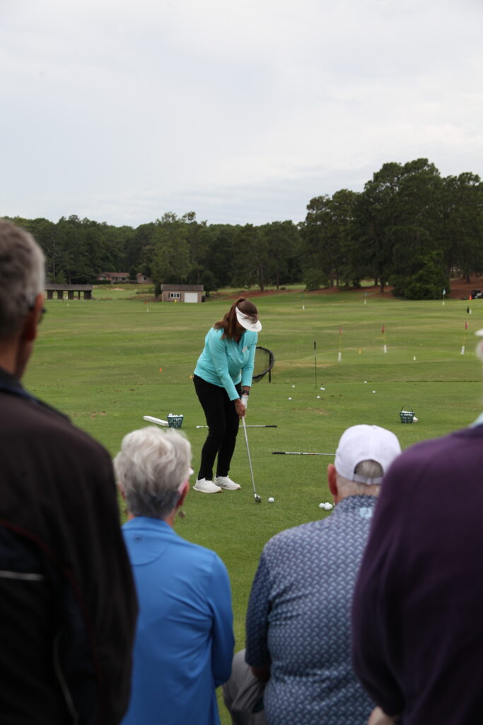 A woman wearing a turquoise jacket and visor prepares to hit a golf ball on a grassy range. In the foreground, several onlookers, including a man in a dark jacket and a woman with gray hair, observe her swing, with trees and practice equipment in the background.