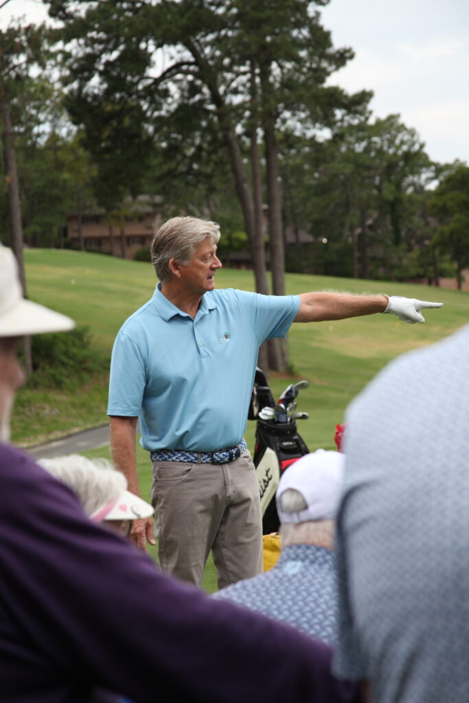 A man in a light blue polo shirt and white gloves gestures with his right hand while addressing a group of people sitting nearby. In the background, a golf course with trees is visible.