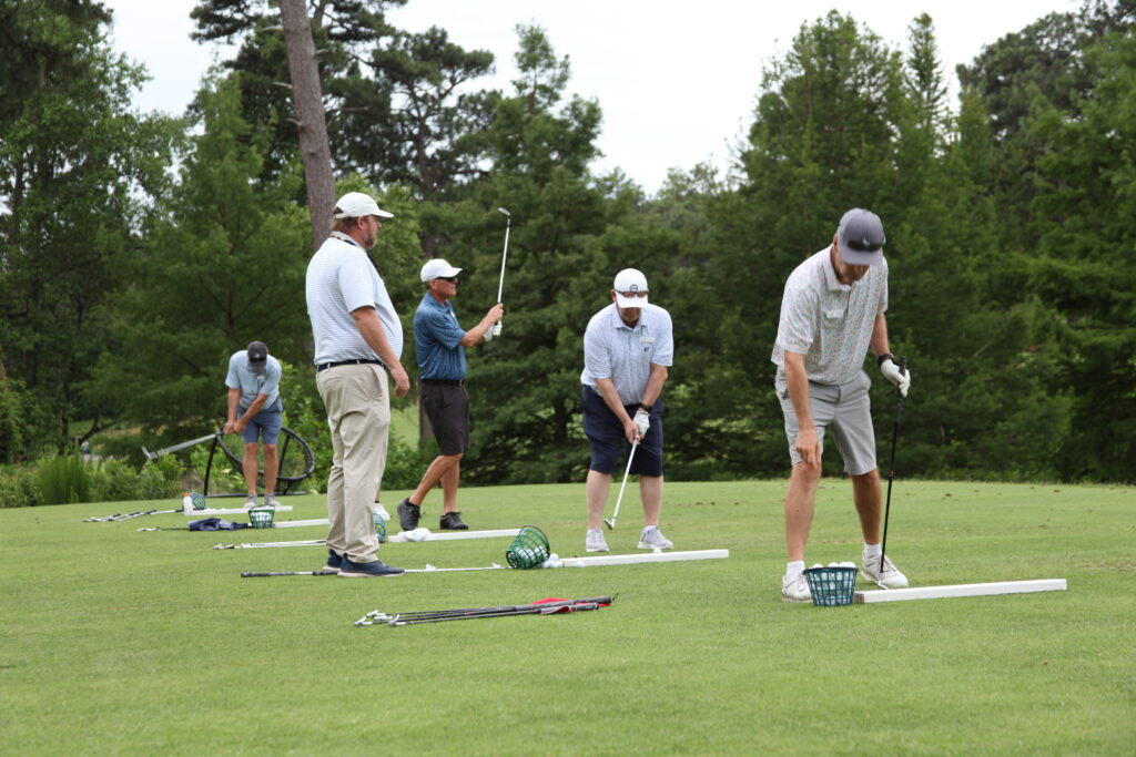 Five golfers on a practice range, each preparing to hit a ball. One golfer in gray pants and a white shirt is addressing the ball, while others are positioned behind him, holding clubs. The background includes trees and grass.