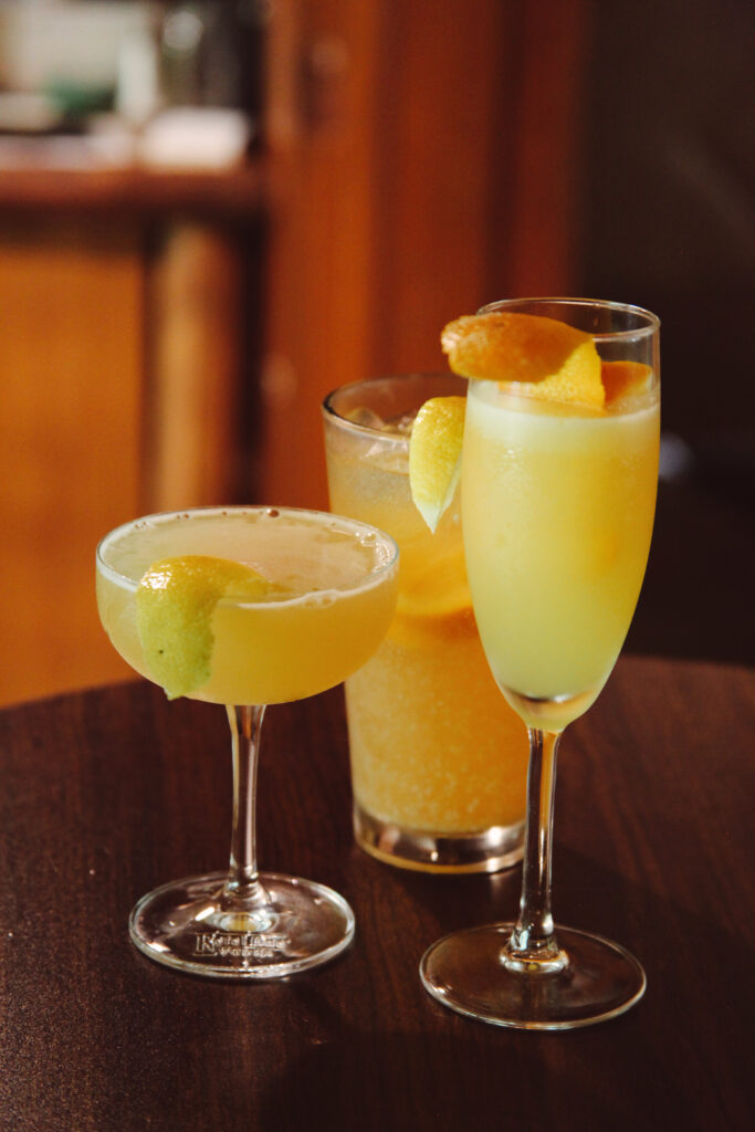 Three colorful cocktails placed on a wooden table. The left drink is a yellow margarita with a lime wedge, the center drink is a light amber beverage, and the right drink is a bright yellow cocktail garnished with orange slices.