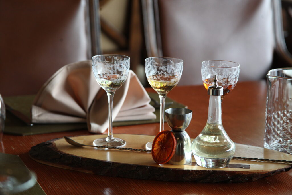 Three ornate glasses containing a light-colored liquid are placed on a wooden serving board. A glass decanter and a metal measure accompany the glasses, along with a round orange slice. In the background, folded napkins are visible on a polished wooden table.