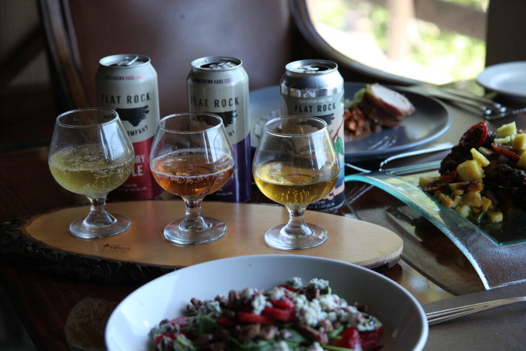 Three small glasses of sparkling cider sit on a wooden tray beside three cans, with a bowl of salad and plates of food on a dining table.