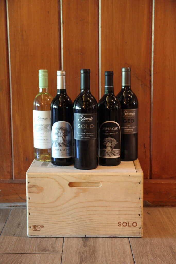 Five wine bottles stand on a wooden crate labeled SOLO against a wood-paneled wall.
