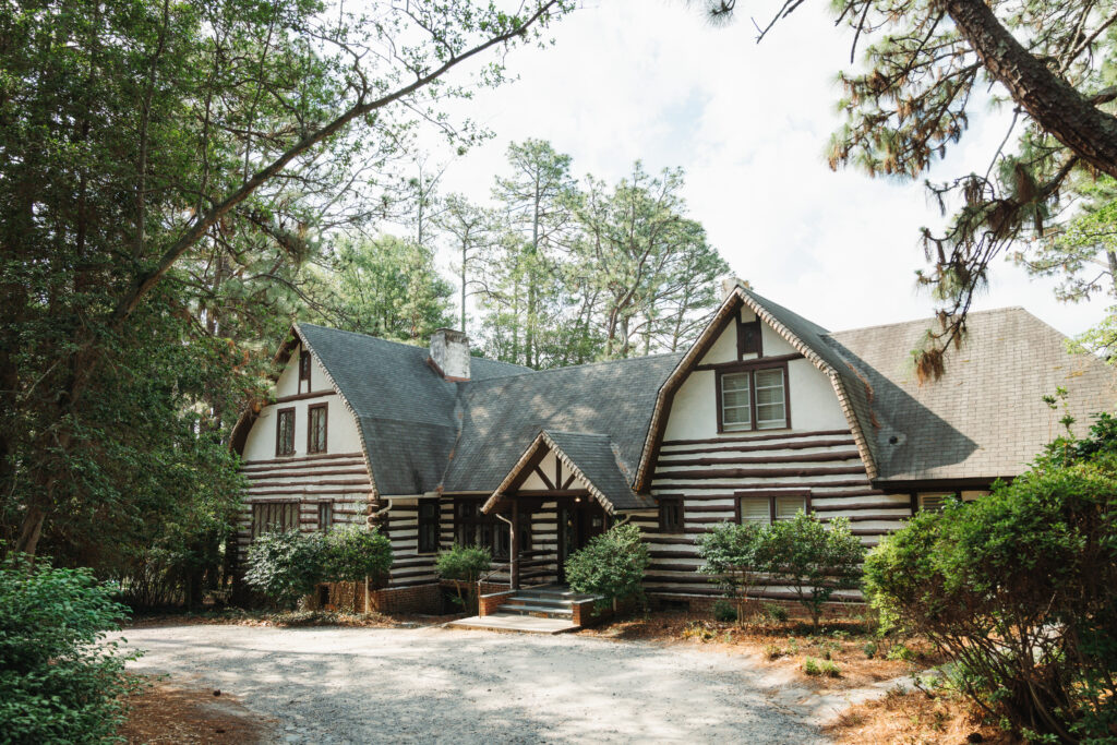 A rustic log cabin-style house with two steep gabled roofs sits among tall trees, with a gravel driveway in front and shrubs around the entrance.