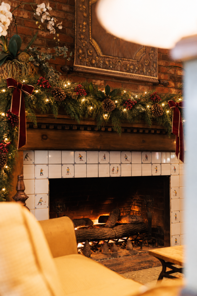 A decorated brick fireplace with a burning fire, garland, red ribbons, and pinecones above a white-tiled surround, with a beige chair in the foreground.