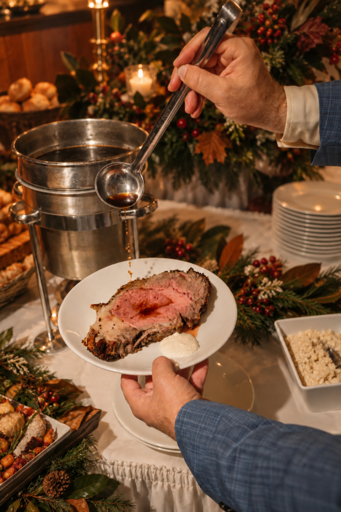 A hand ladles sauce onto a sliced roast beef on a white plate, beside a metal serving urn and holiday greenery on a buffet table.
