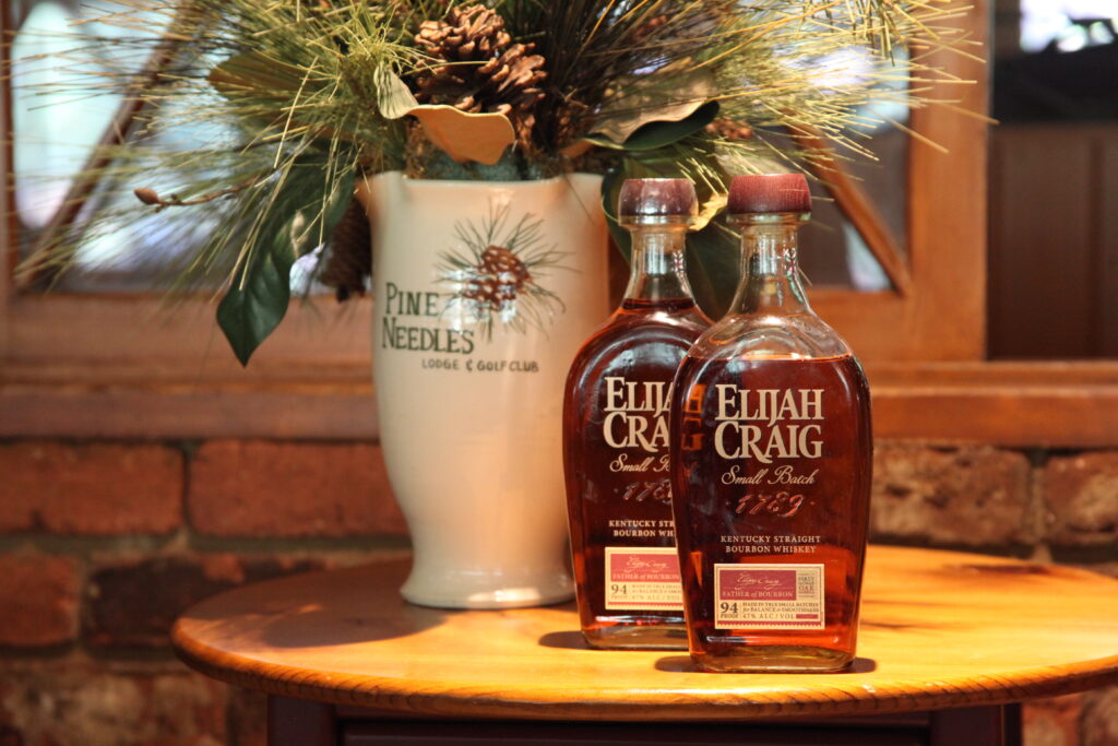 Two Elijah Craig bourbon bottles stand on a round wooden table beside a white Pine Needles vase holding greenery and pinecones.