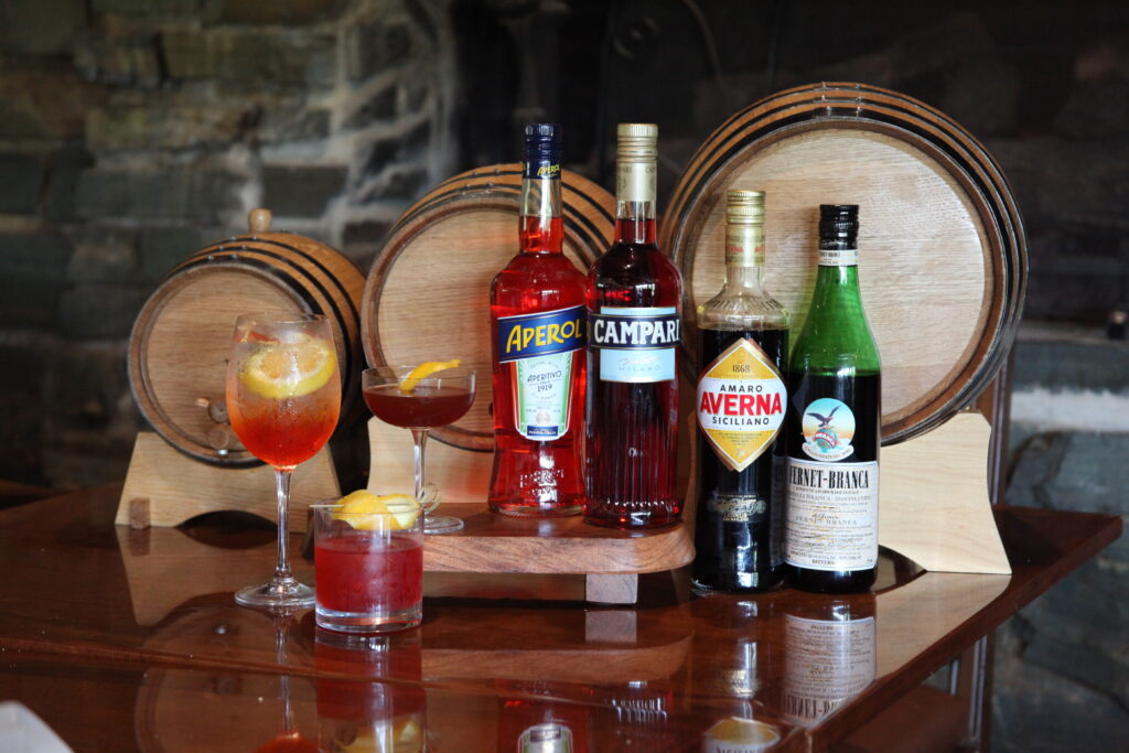 A display of Aperol and Campari bottles with three cocktail glasses and lemon slices on a polished wooden table, arranged in front of round wooden barrels.