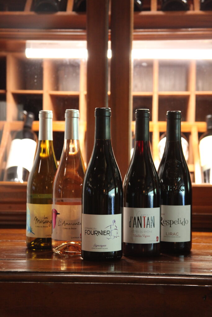 Five wine bottles stand on a wooden counter in a dimly lit wine cellar, with shelves and lights in the background.