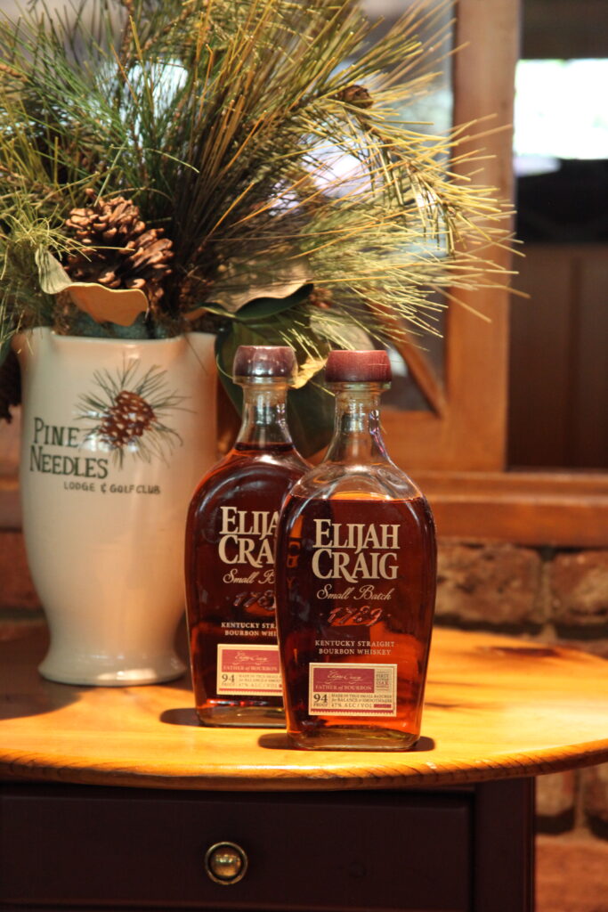 Two Elijah Craig whiskey bottles sit on a wooden table beside a pine needle arrangement in a white vase.